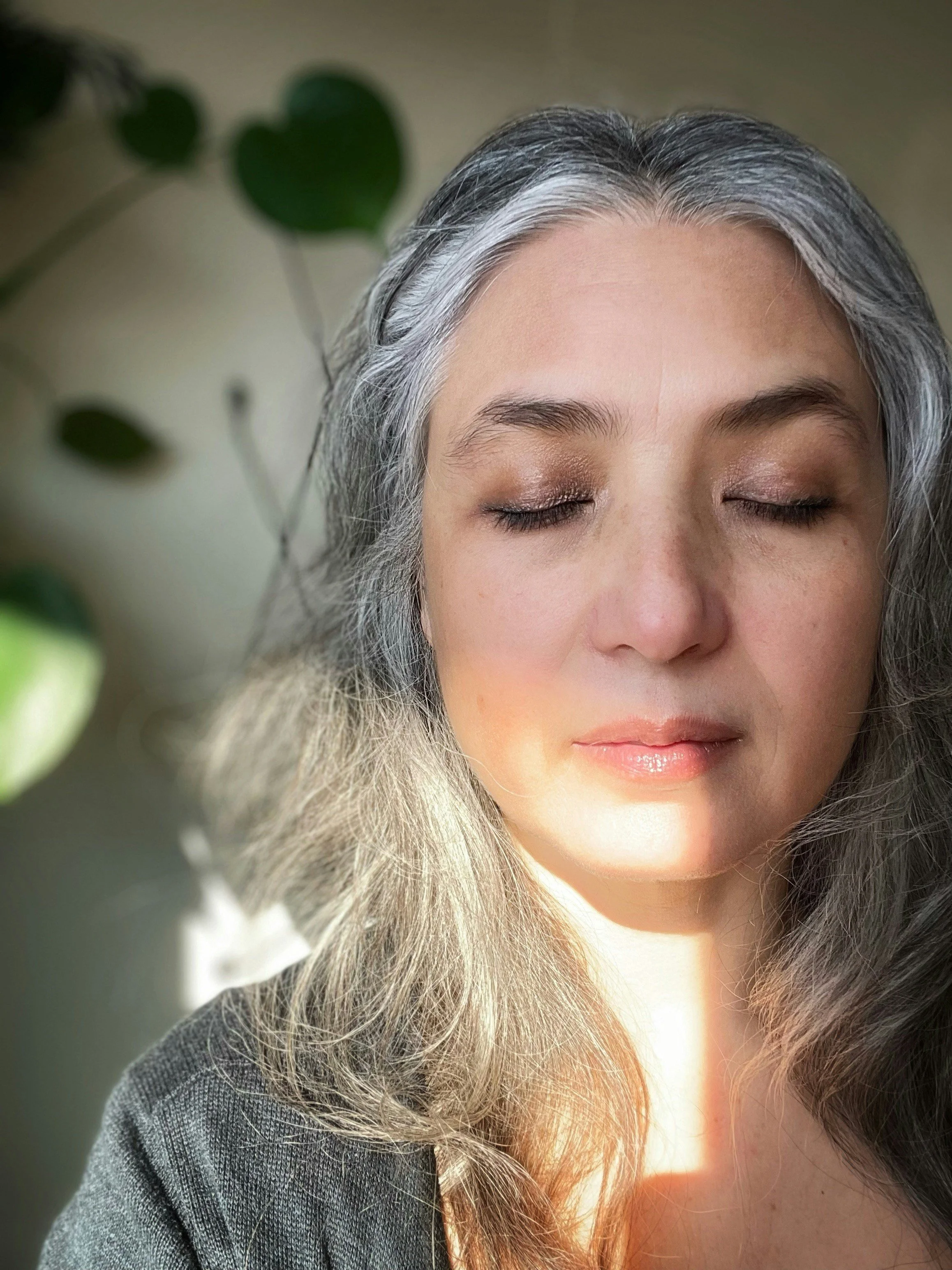 Close-up of a woman's face with long, wavy gray hair, closed eyes, and soft lighting, with greenery in the background.