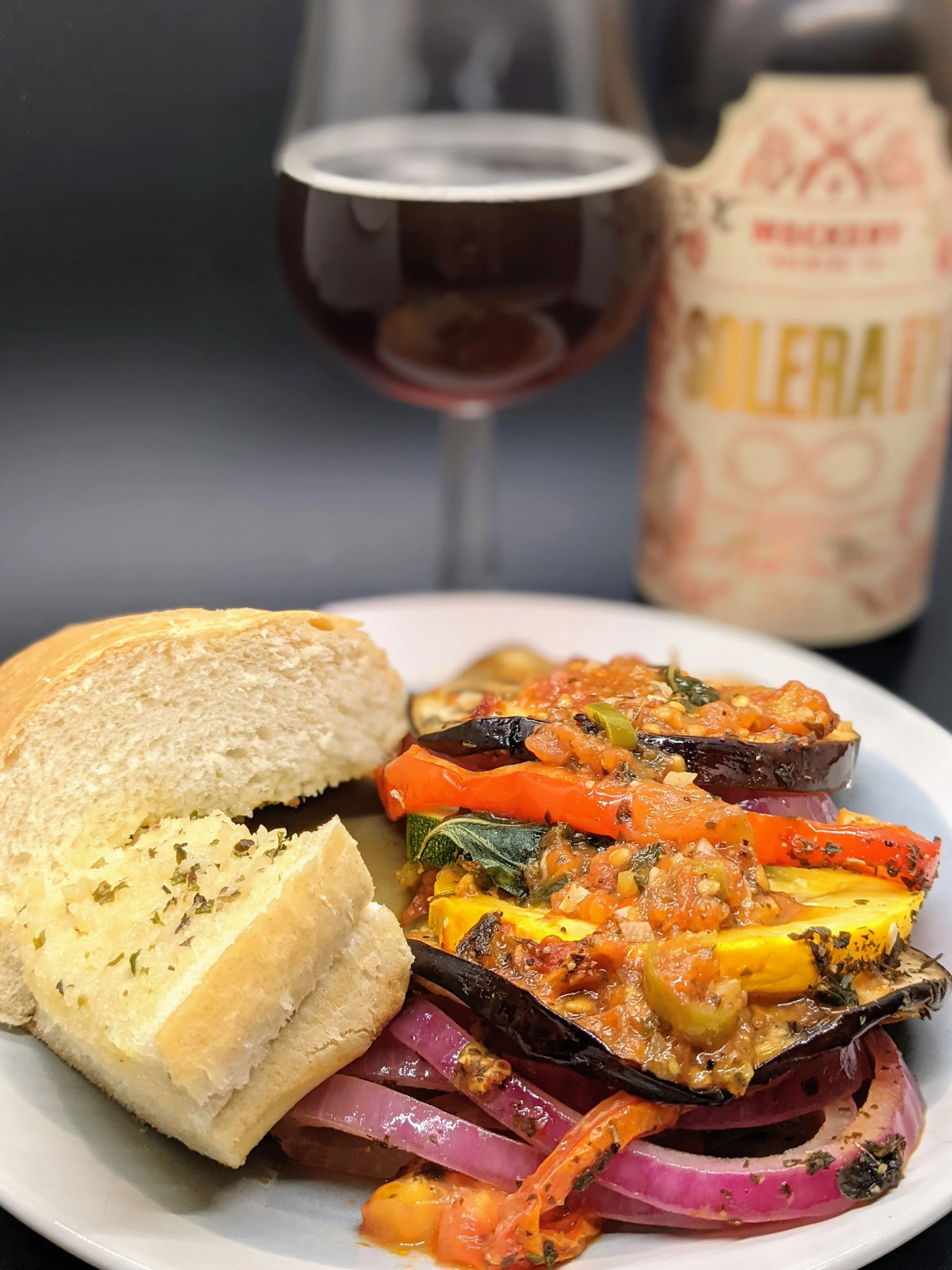 Image of a delicious plate of Ratatouille served with a slice of garlic bread, paired with a craft beer in the background. The Ratatouille is a vegan dish featuring a medley of fresh vegetables and spices, while the beer provides a fruity and acidic