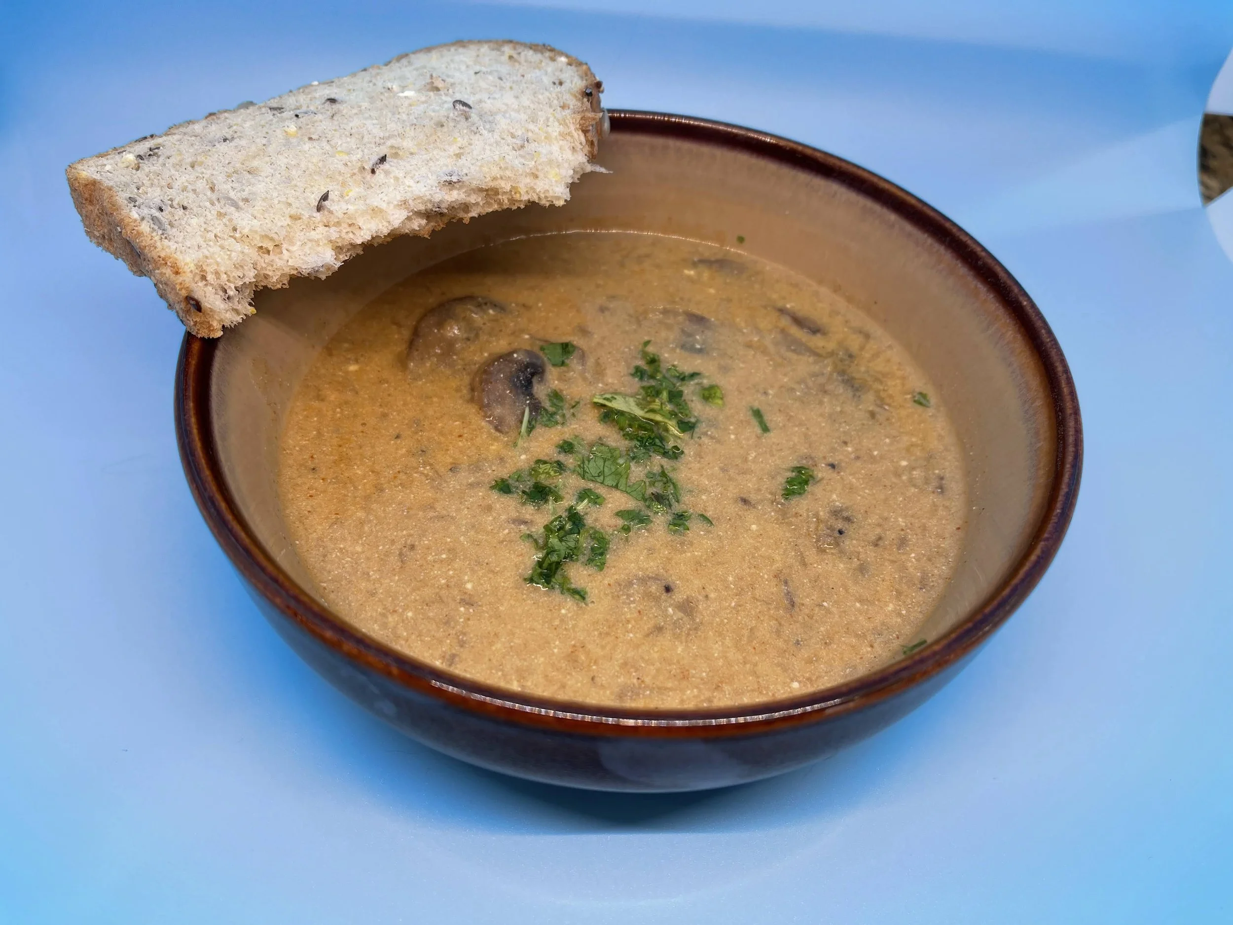 A bowl of savory mushroom soup, with a slice of toast resting on the edge of the bowl, surrounded by sliced mushrooms and parsley leaves