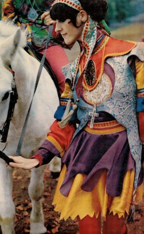A person wearing layered, medieval-inspired, multi-colored garb stands beside a white horse, the clothing is a riot of bright colors and includes an elaborate headband, tooled leather-seeming tunic, hose, and jagged and notched hemlines