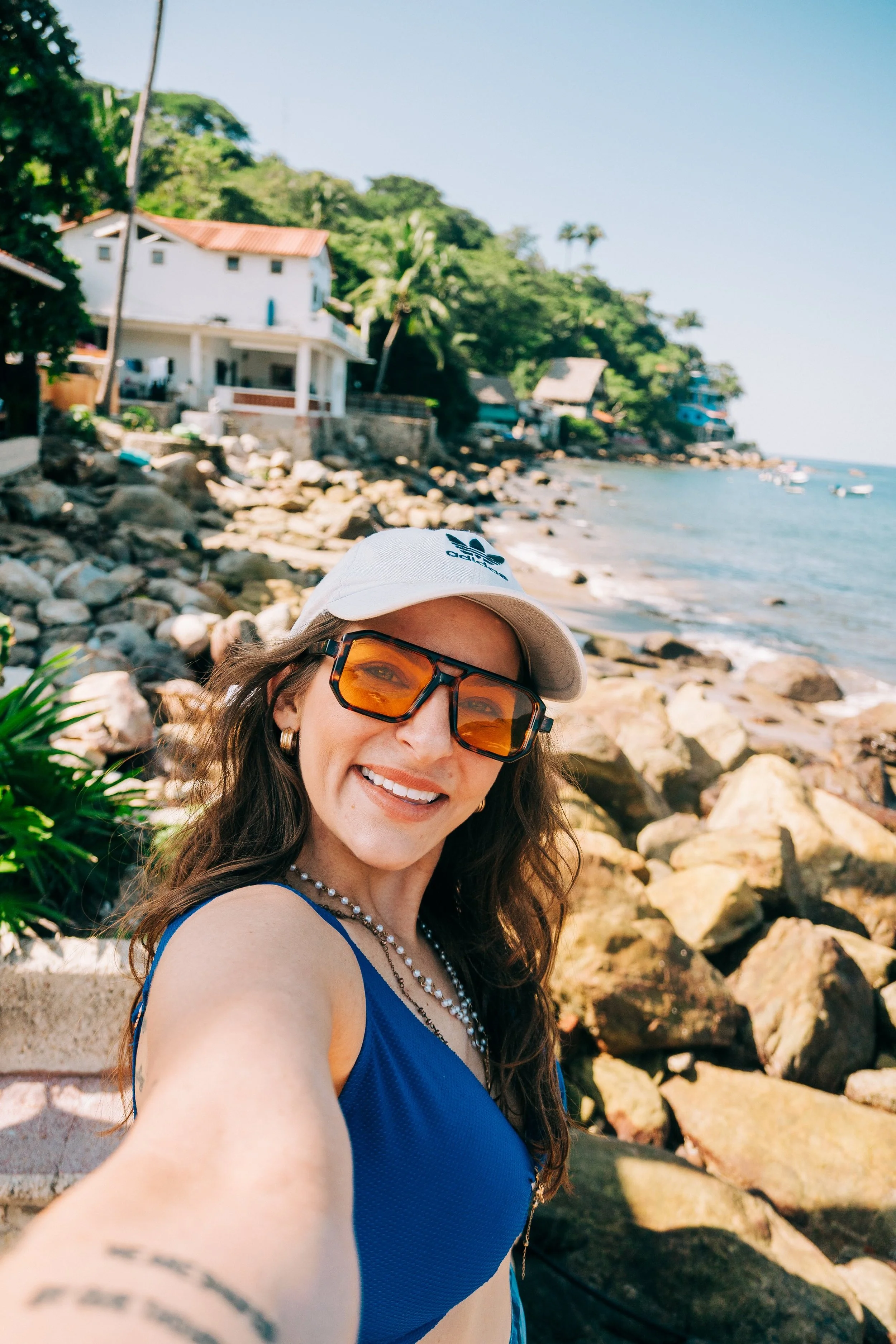 Katie Coffee in Puerto Vallarta smiling at a rocky beach, wearing sunglasses, a white cap, and a blue top, nashville travel wellness photographer