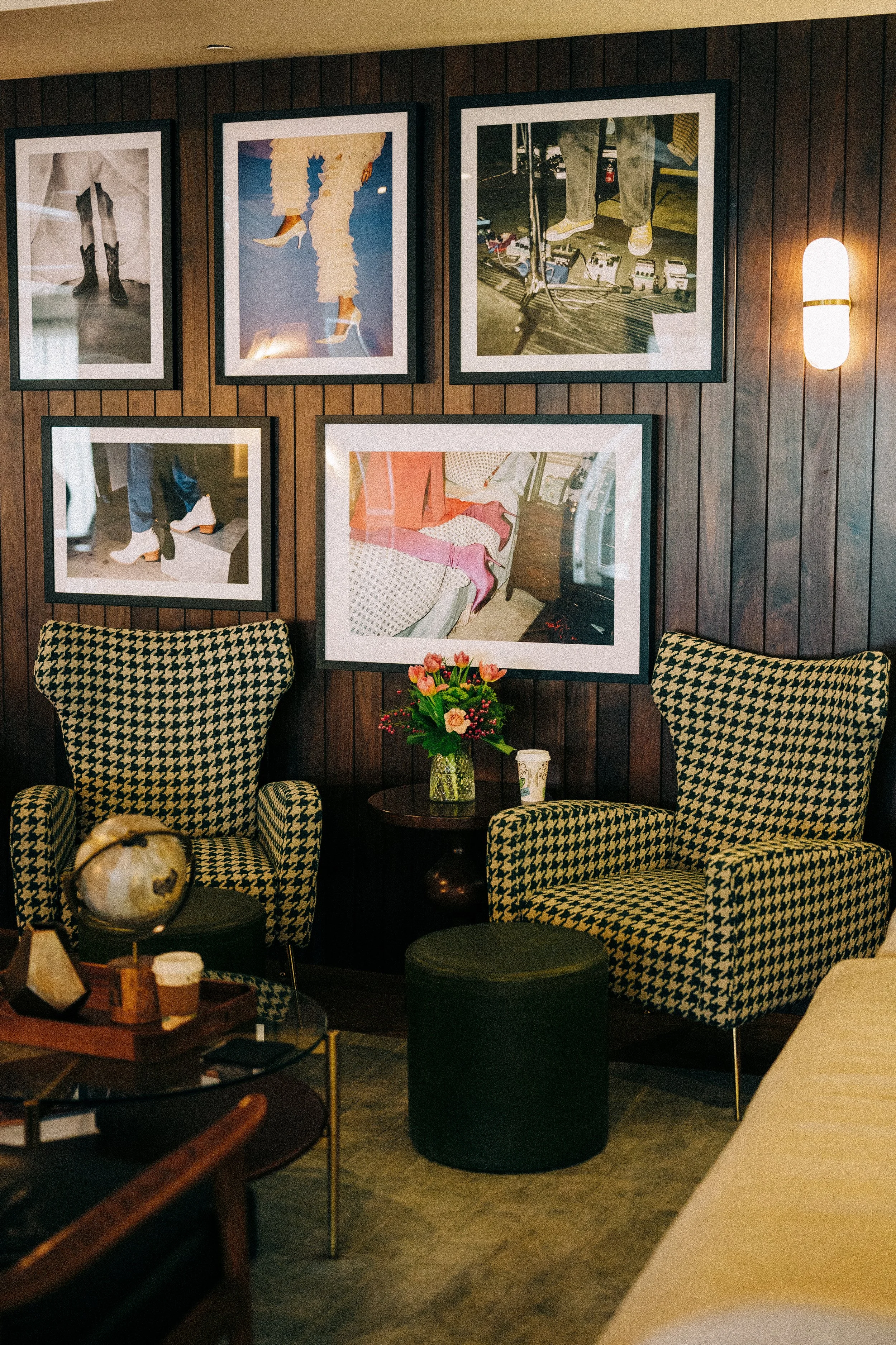 A cozy seating area with two houndstooth armchairs, a small wooden table with a flower vase, framed photographs on a wooden wall, and a soft wall sconce light.