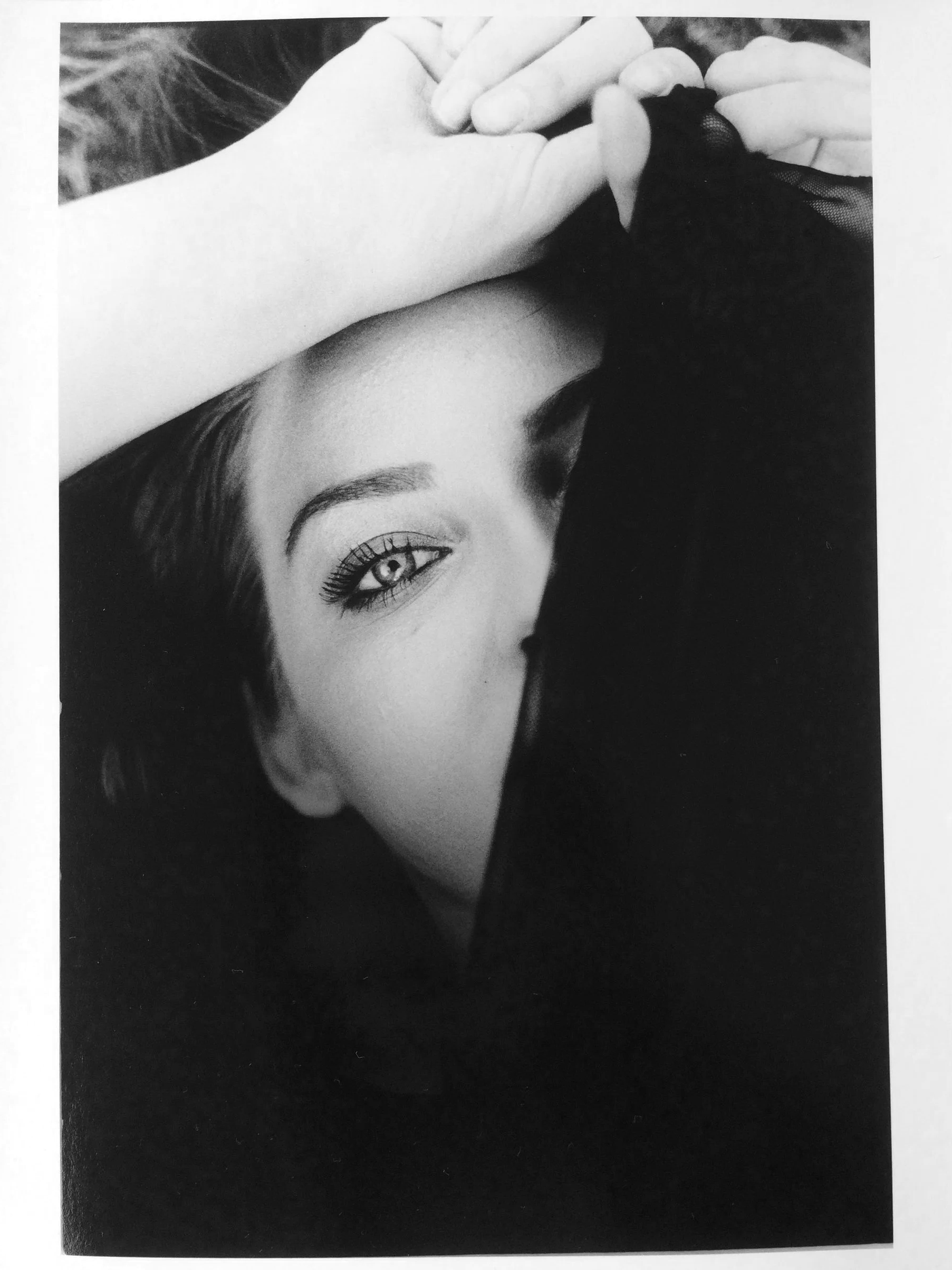 Black and white photograph of a woman lying down, partially covered by a black piece of cloth, with one eye visible, looking directly at the camera.