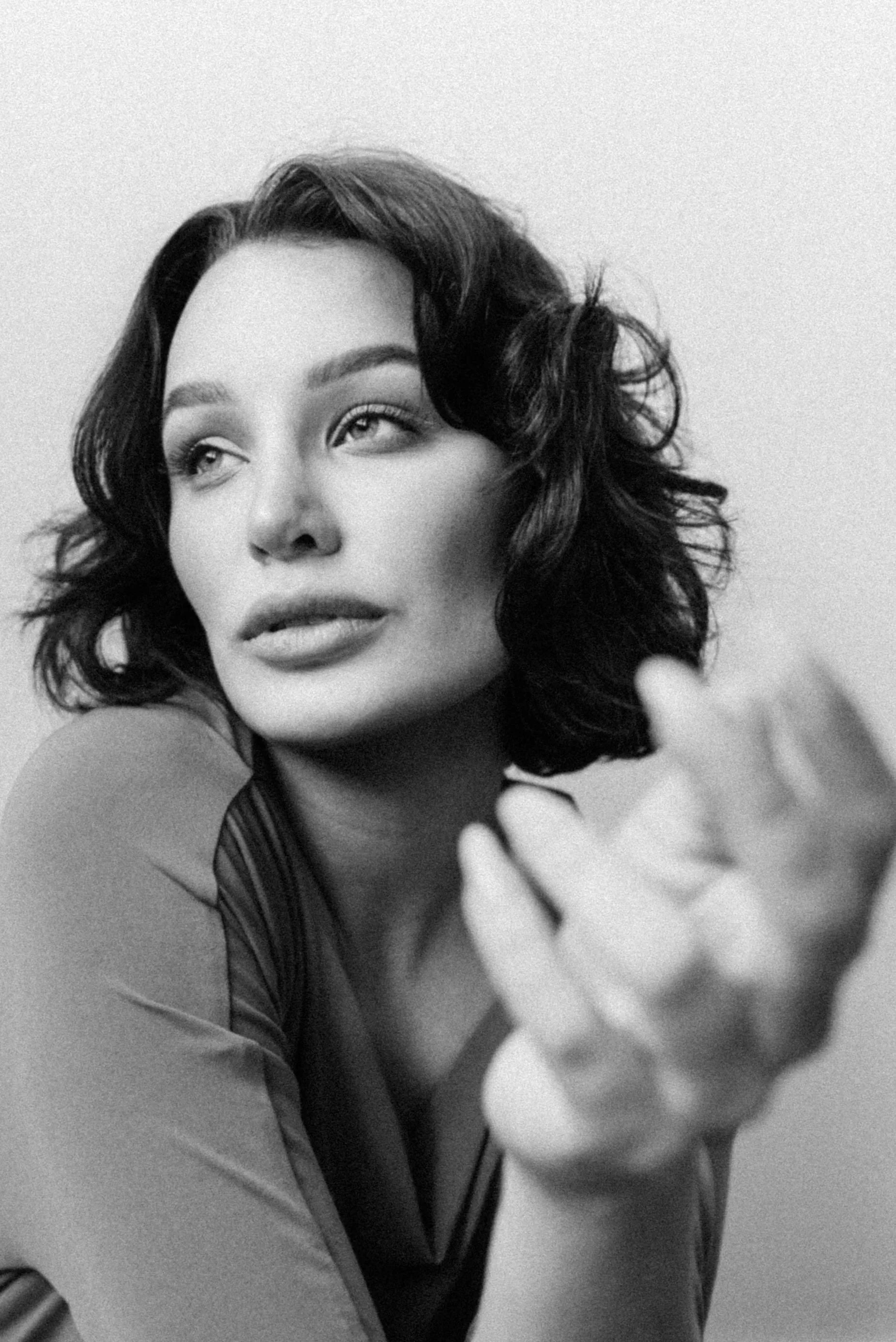 Black and white portrait of a woman with wavy shoulder-length hair, looking slightly to her right, with her hand extended towards the camera.