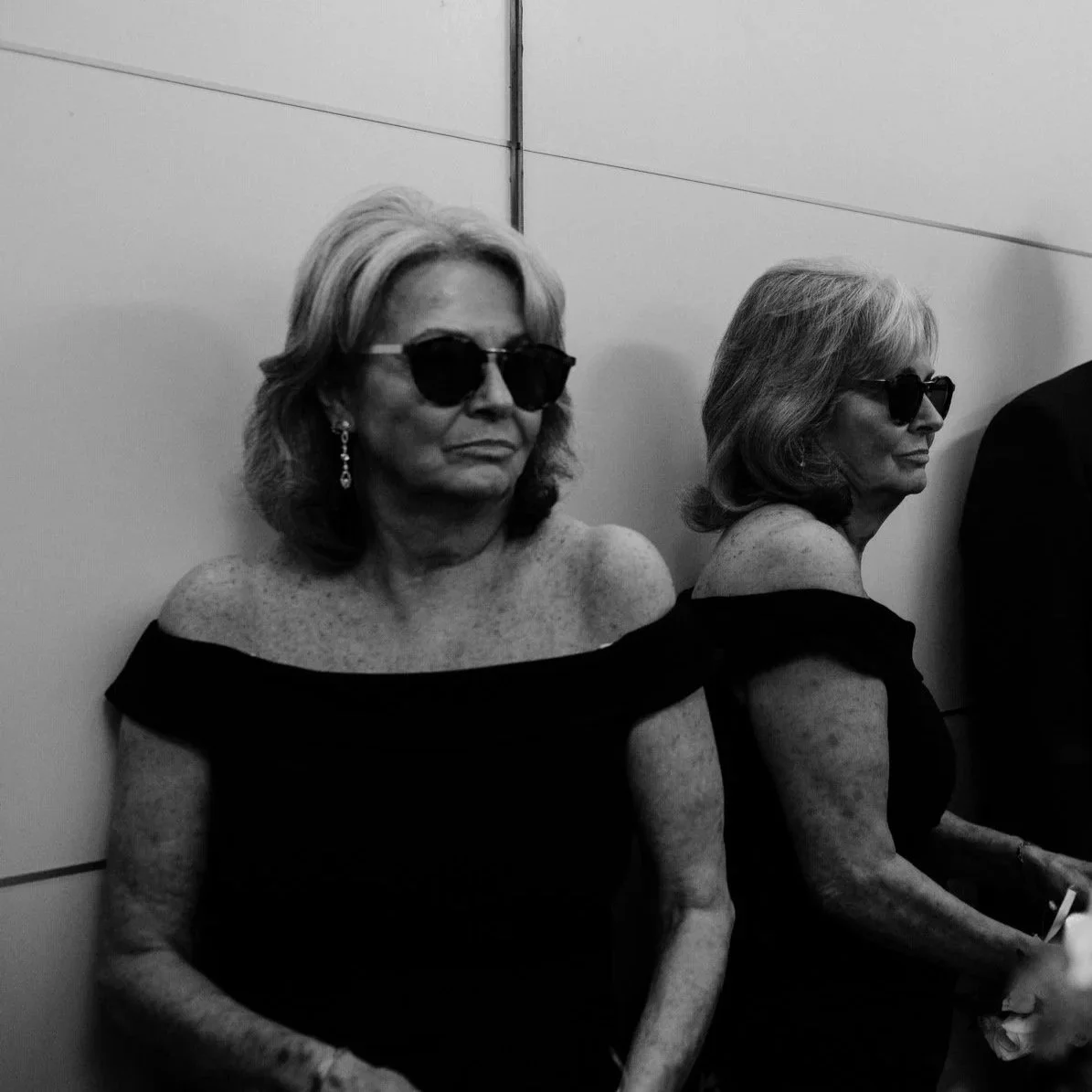 Two elderly women wearing sunglasses, black off-shoulder dresses, with short hair, sitting against a plain wall.