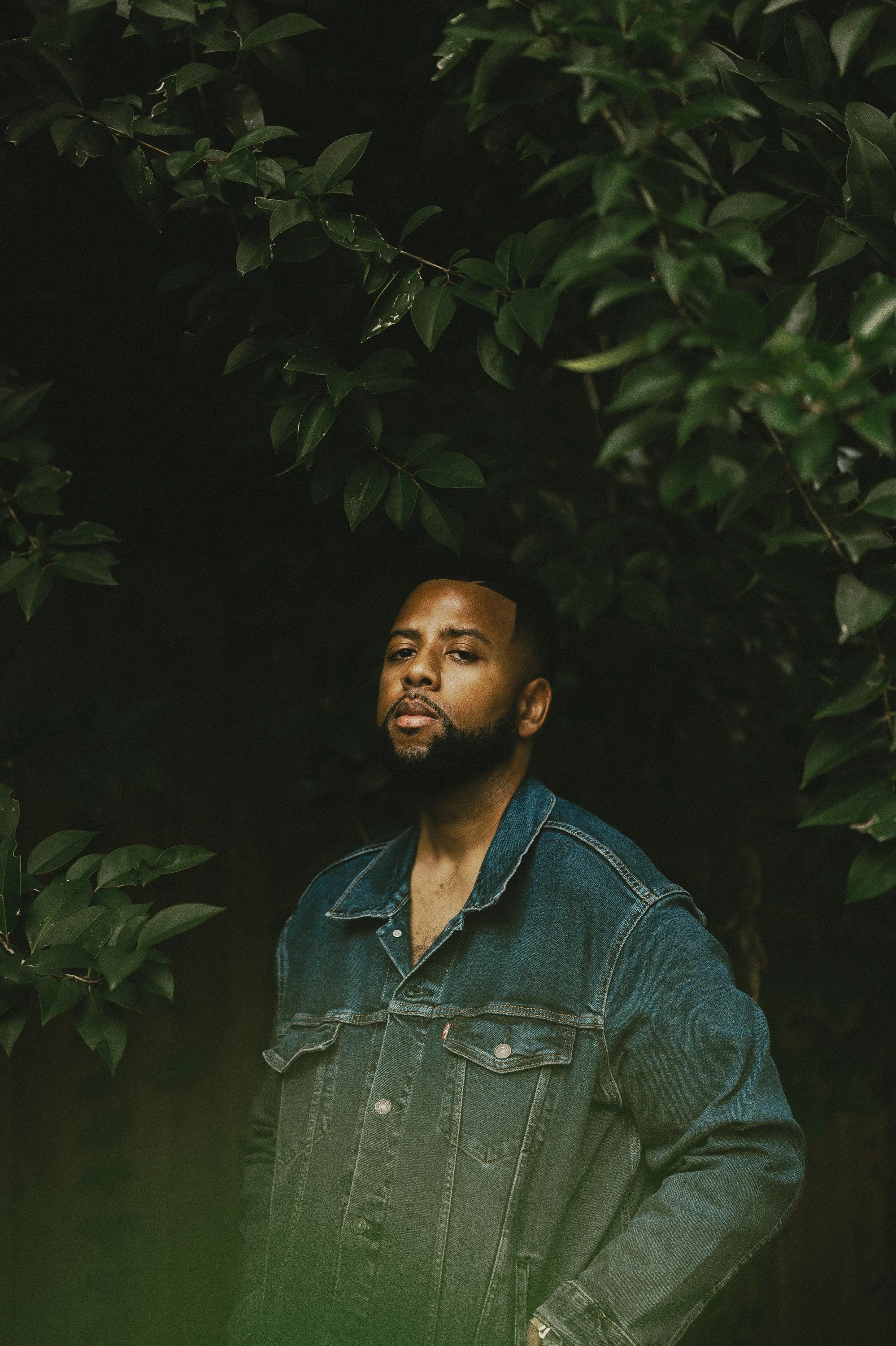 A man with a beard wearing a denim jacket standing among green leaves.