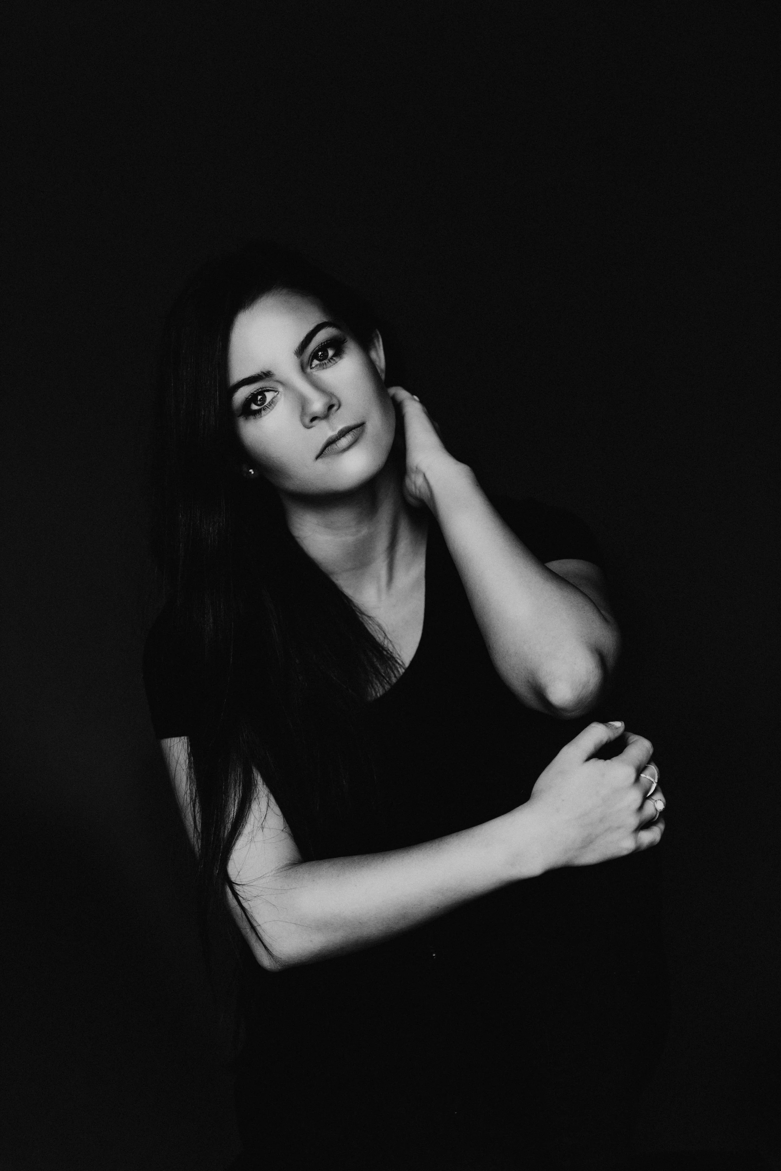 Black and white portrait of a woman with long dark hair, wearing a black top, looking at the camera with a relaxed pose, one hand touching her neck and the other resting on her arm.