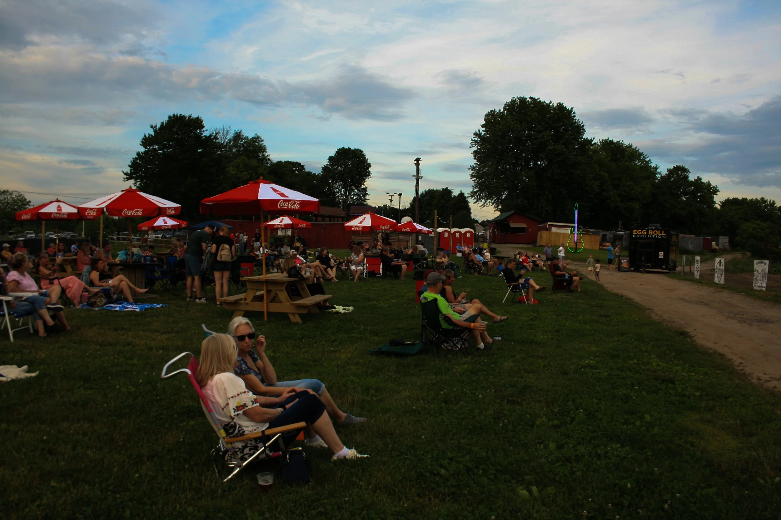A crowd of people sitting on lawn chairs and picnic blankets outdoors during evening, with red Coca-Cola umbrellas, a stage with neon guitar sign, and trees in the background.