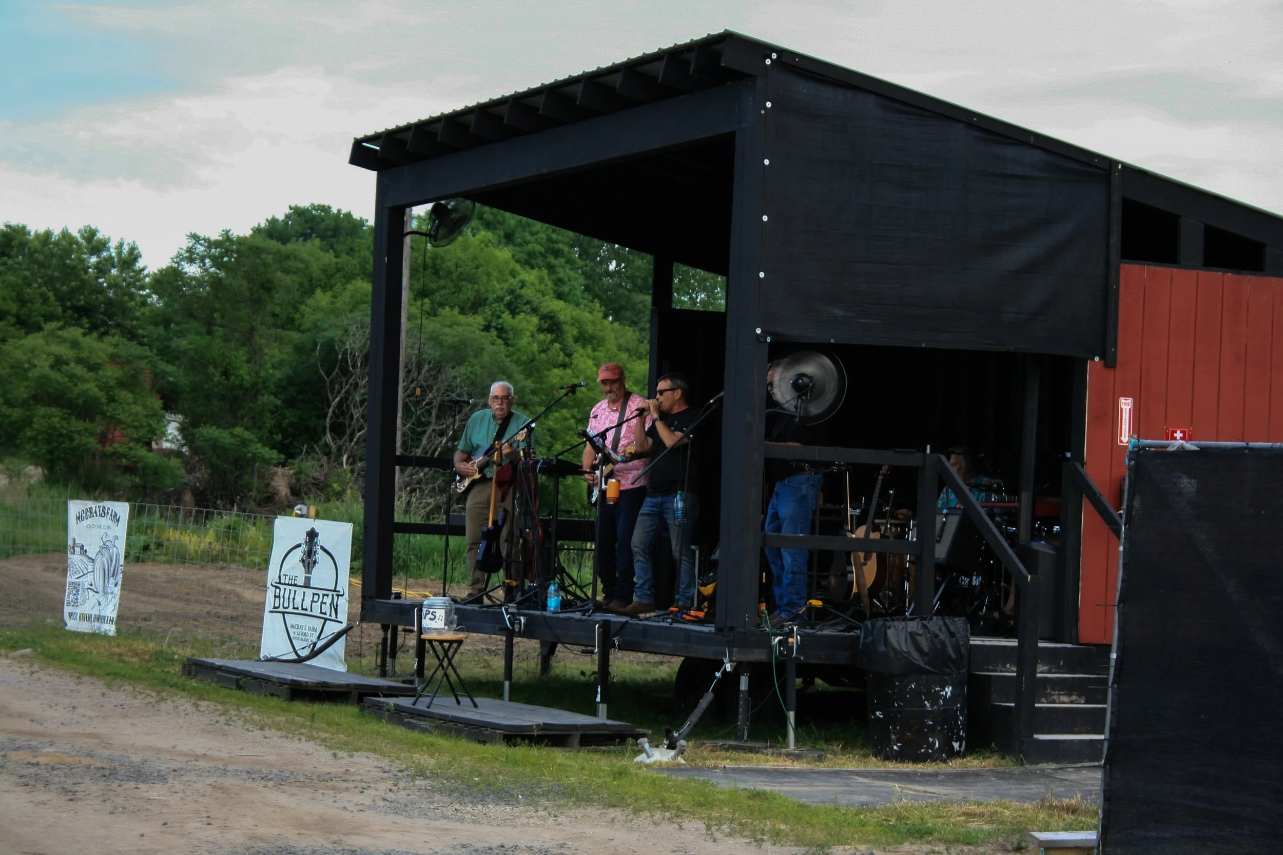 A small outdoor stage with three musicians performing in front of a green, wooded background. The musicians include an older man with glasses playing a guitar, a man in a pink shirt and hat, and a man with glasses singing into a microphone. There are