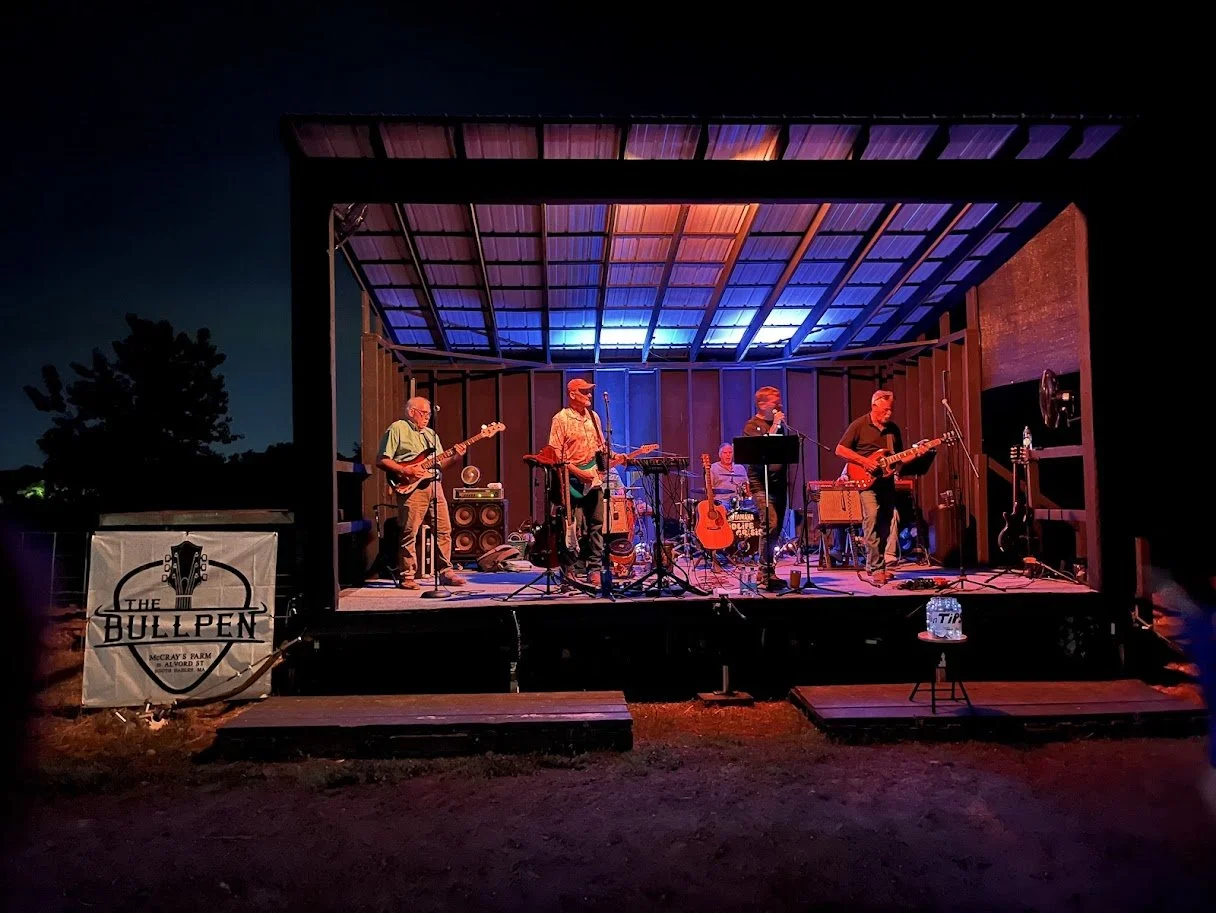 A band performing live on an outdoor stage at night, illuminated by colorful lights, at McCray's Farm with a sign that reads "The Bullpen".