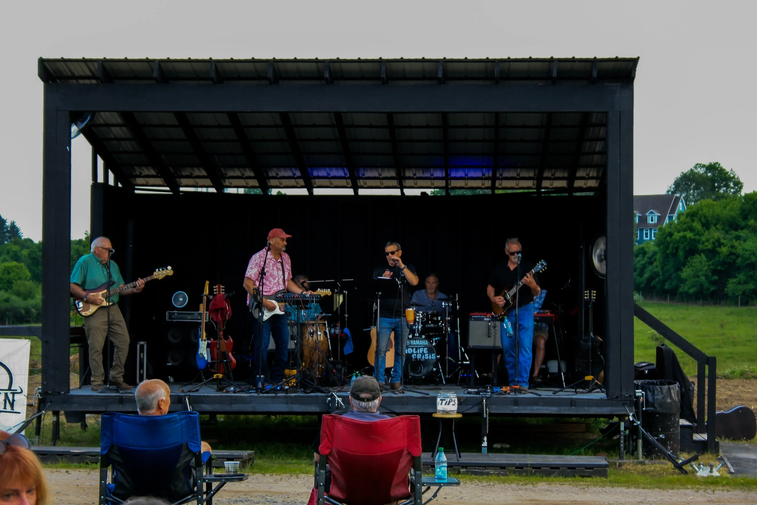A live outdoor band performance on a stage with five musicians, including guitarists and a drummer, during daytime, with audience members seated in chairs in front of the stage.