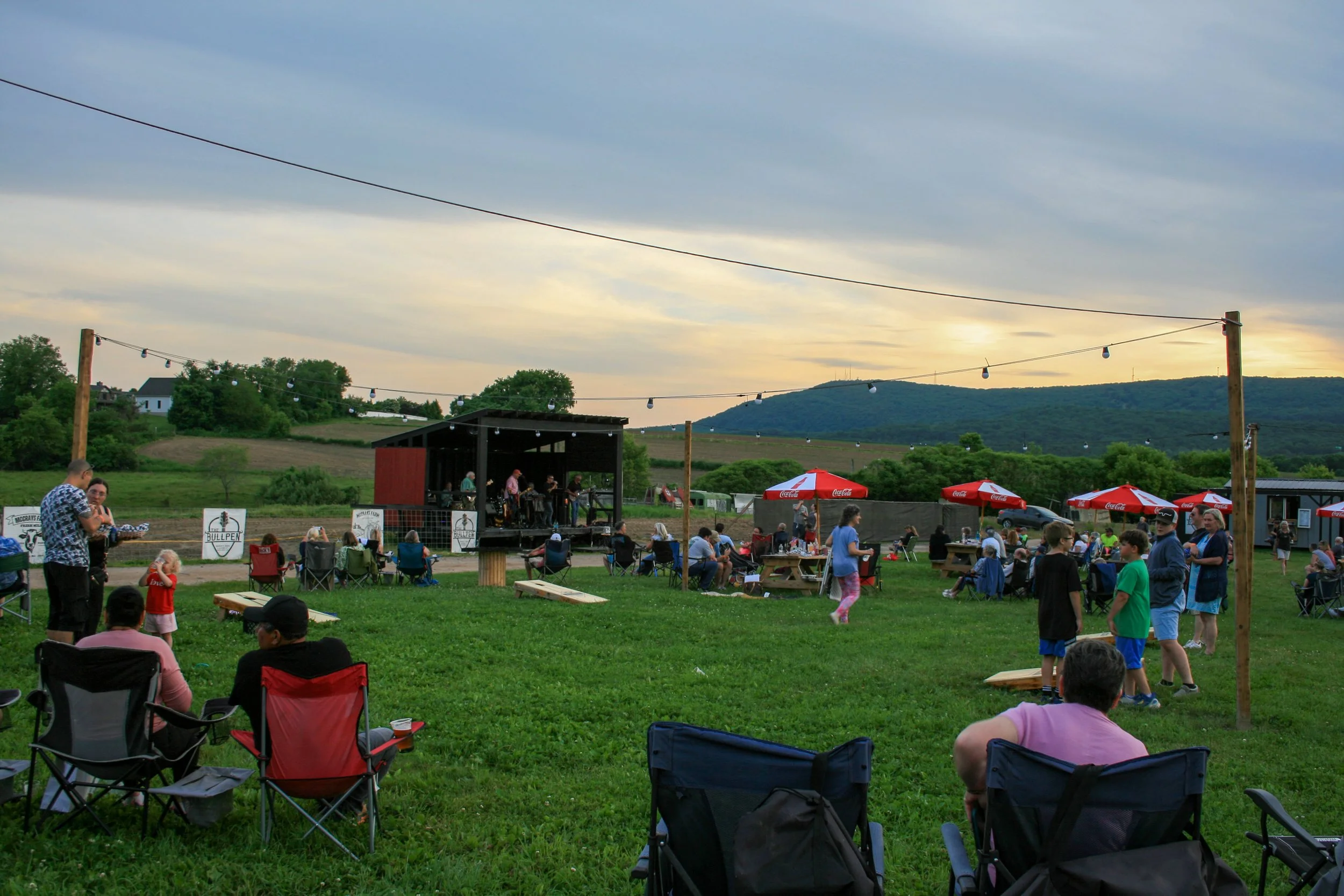 Outdoor community event with people sitting on chairs and standing around, a small stage with a band, string lights, red umbrellas, and a rural landscape with hills and trees in the background during sunset.