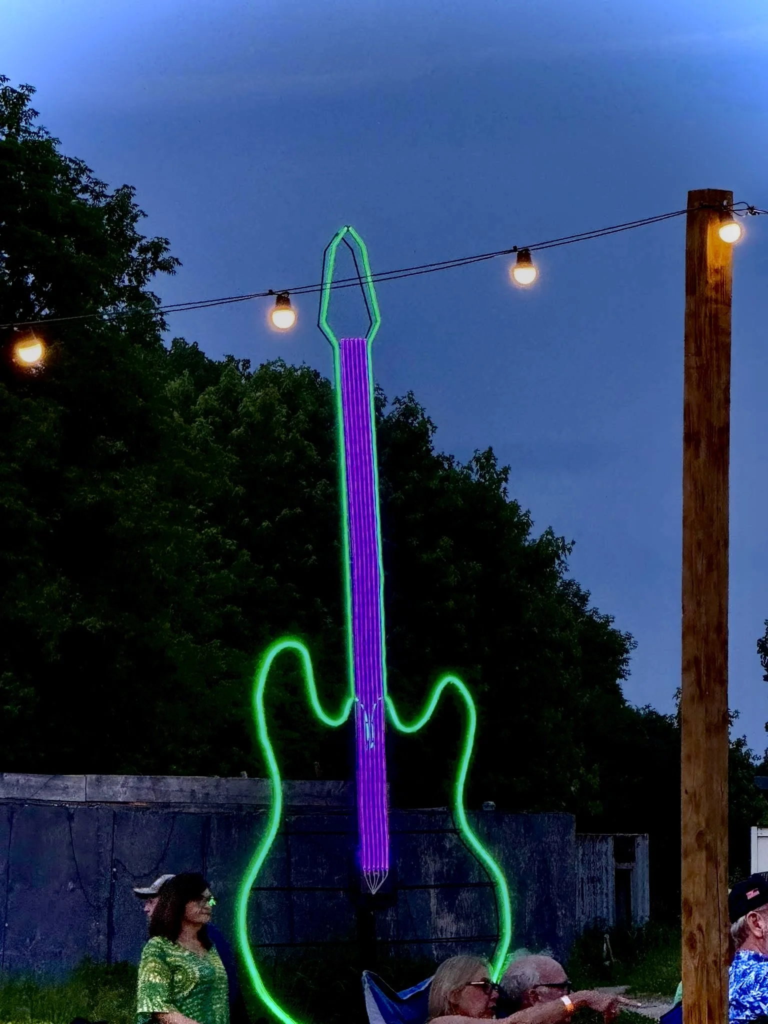 Neon light installation in the shape of a fork and spoon, hanging above a group of people outdoors during dusk, with trees and a wooden pole in the background.