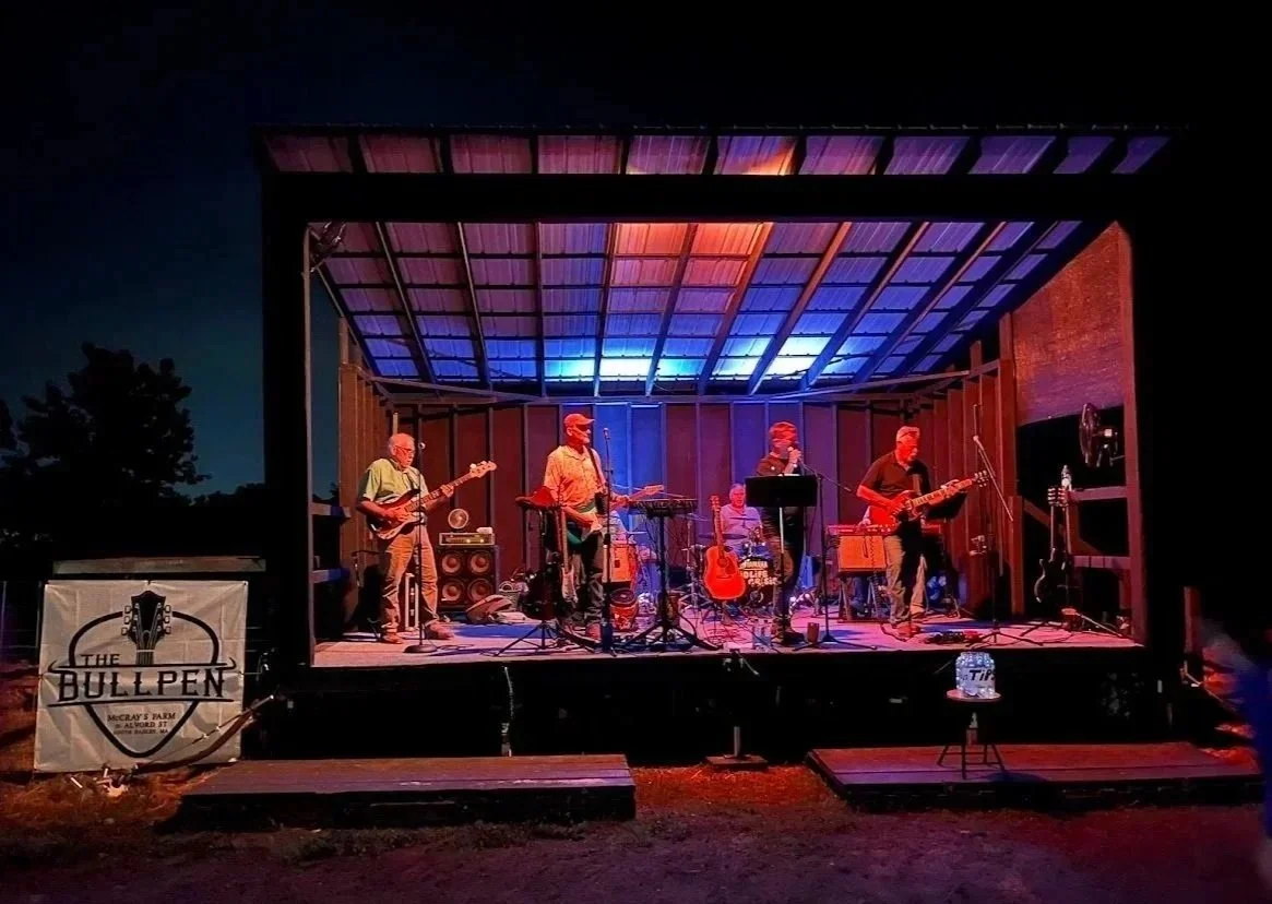 A live band performing on an outdoor stage at night with colorful blue and purple lighting, featuring five musicians playing guitars, drums, and keyboard.