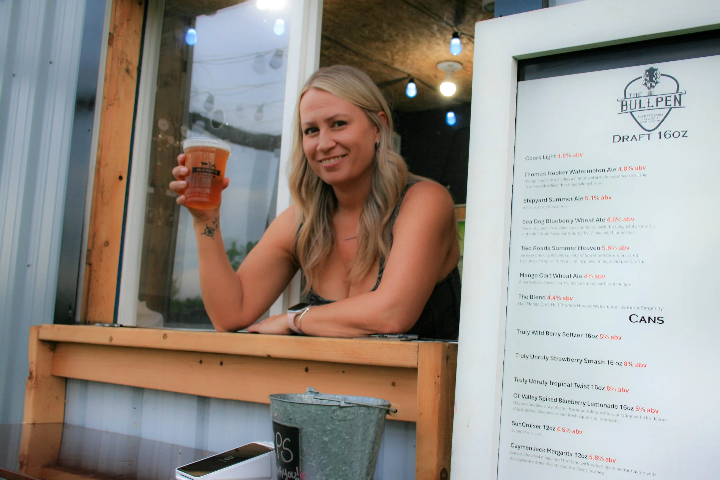 A woman with blonde hair holding a plastic cup of beer at a bar or outdoor drink stand, smiling towards the camera. The background includes a wooden and metal structure and a menu board with various beer options.