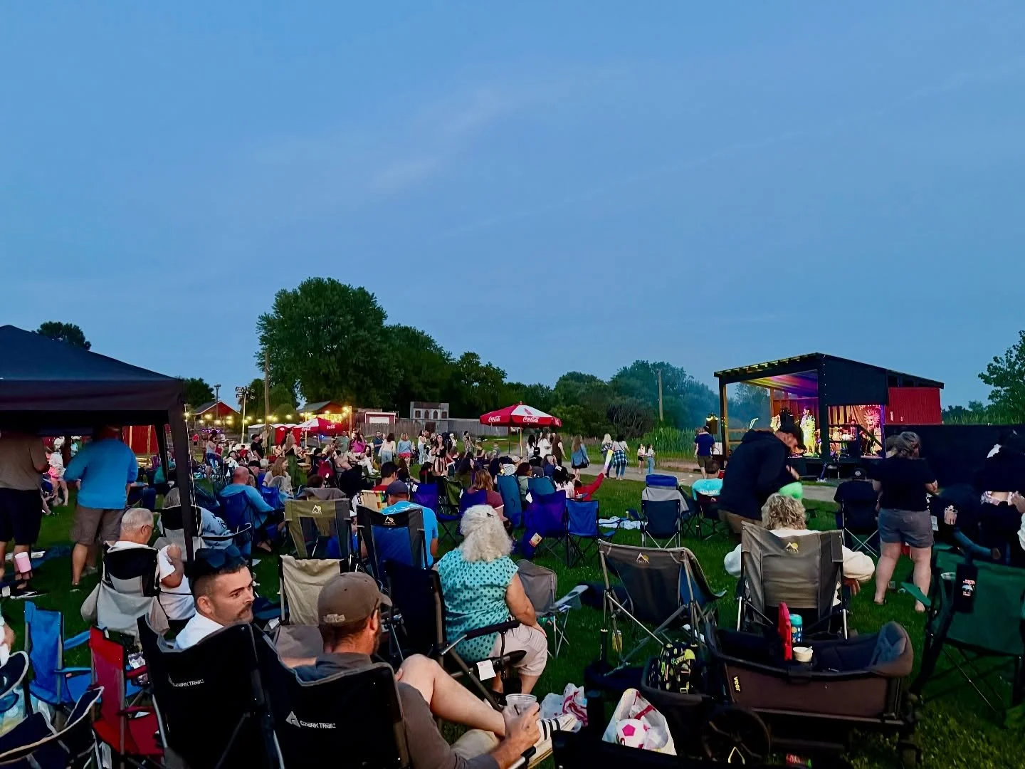 Outdoor concert scene with people sitting on chairs and standing, watching a stage with performers, under dusk sky with trees and umbrellas in the background.