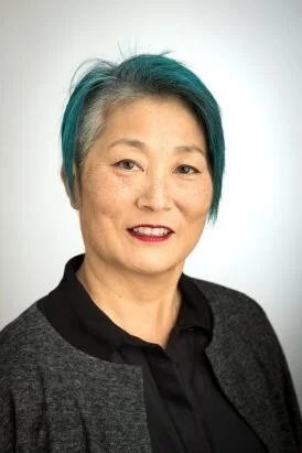A photo of Lisa Nakamura, a woman with short gray hair, dark lipstick, and a black and gray shirt