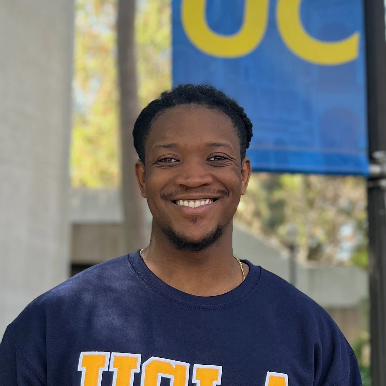 A photo of Ngozi Harrison, a Black man, standing outside under a small banner with the letters "UC." He has short hair, a kind smile, dark brown eyes, and a UCLA sweater on.