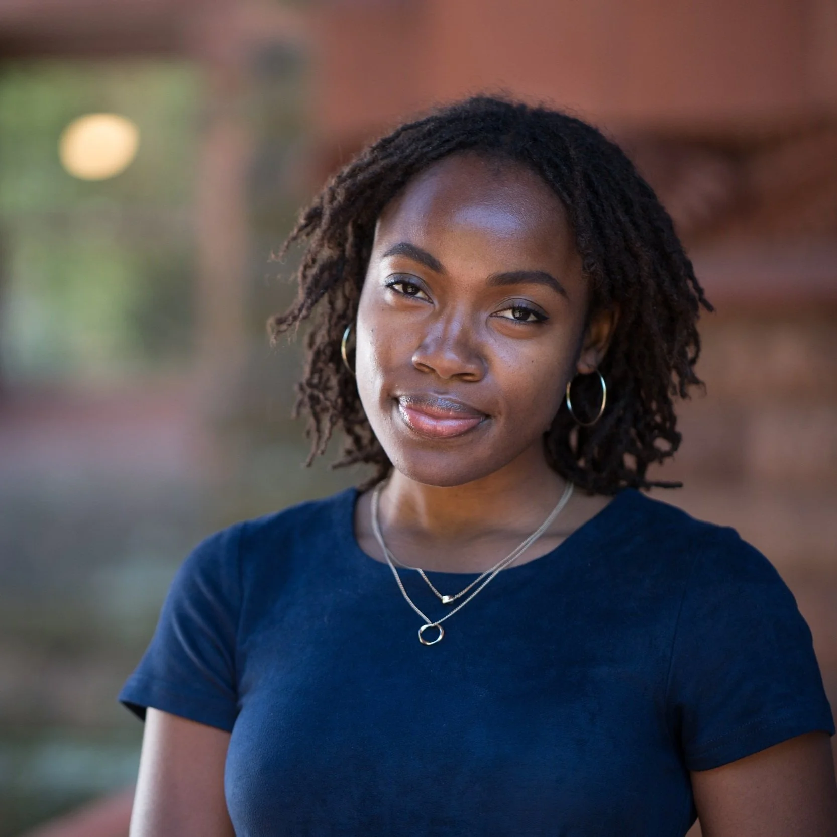 Cienna Davis, a Black woman, stands in front of a blurred brown interior wall, wearing two small silver necklaces and a navy blue t-shirt. She has a gentle smile and a shoulder length bob haircut.