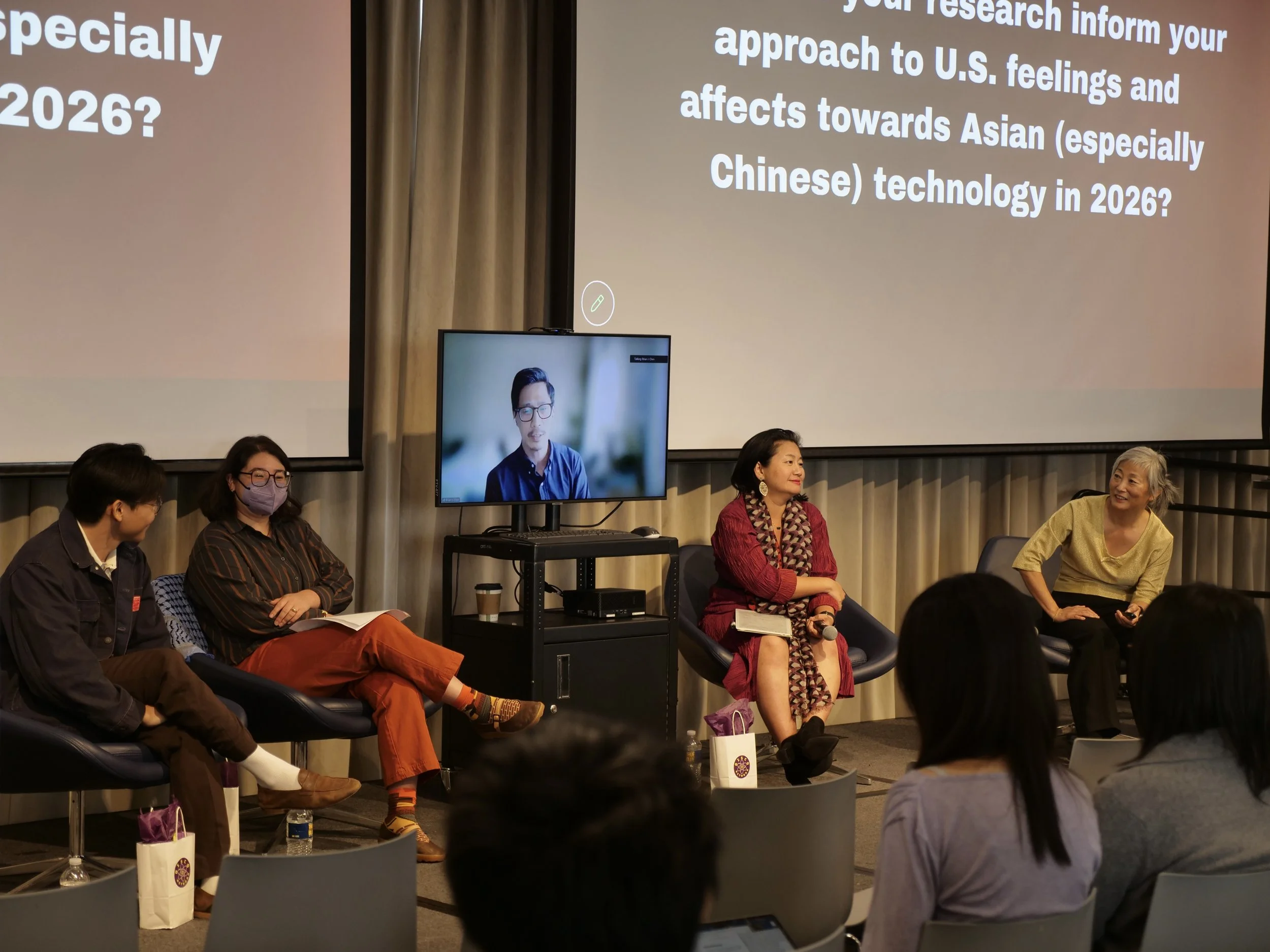 Panel discussion with four participants, including one appearing on a monitor, seated before projection screens while an audience watches.