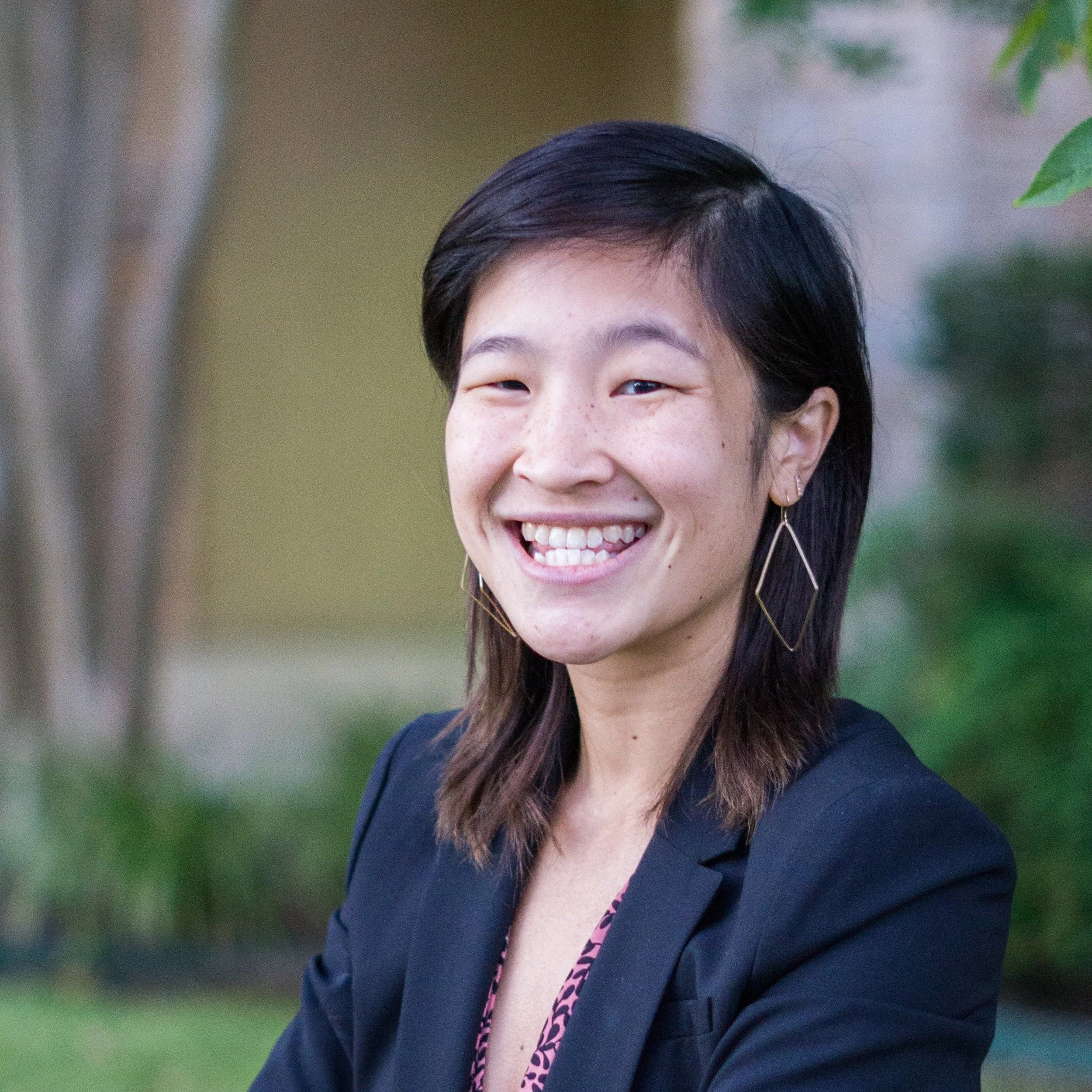 A headshot of Michelle Lee, an East Asian woman, standing outside in front of some greenery. She has a navy blazer on, shoulder length hair, dangling diamond-shaped earrings, and a large smile.