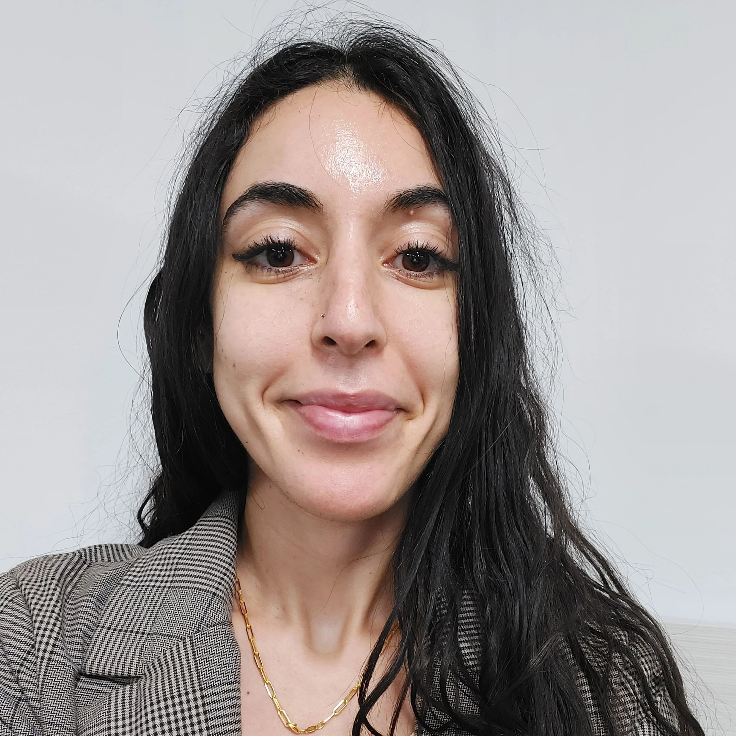A photo of Sara Abdulla, a woman with long dark hair and a gray blazer standing against a lighter background, smiling.
