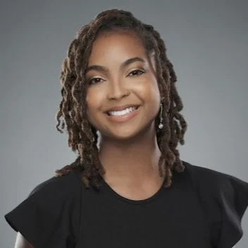 A photo of Nina-Simone Edwards, a Black woman with a bright smile and brown hair reaching to the collar of her shirt. She's wearing a black blouse with slightly ruffled short sleeves.
