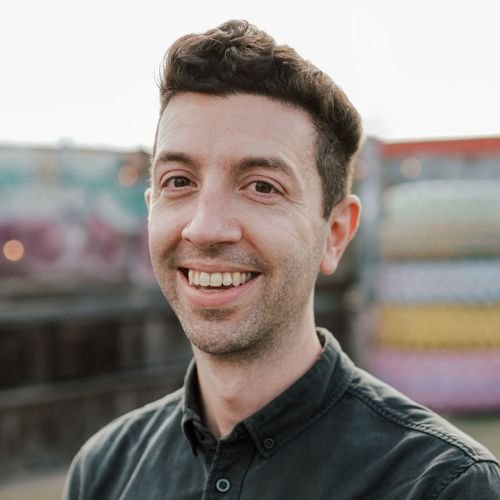 A portrait of Tim Newman, a white man, standing outside against a blurred, colorful wall. He has a large smile, brown eyes, short slightly curly hair, and wears a dark button down.