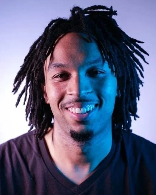 A photo of Glenn Cantave, a Black man with shoulder length hair, a goatee, and a smile in bluish lighting