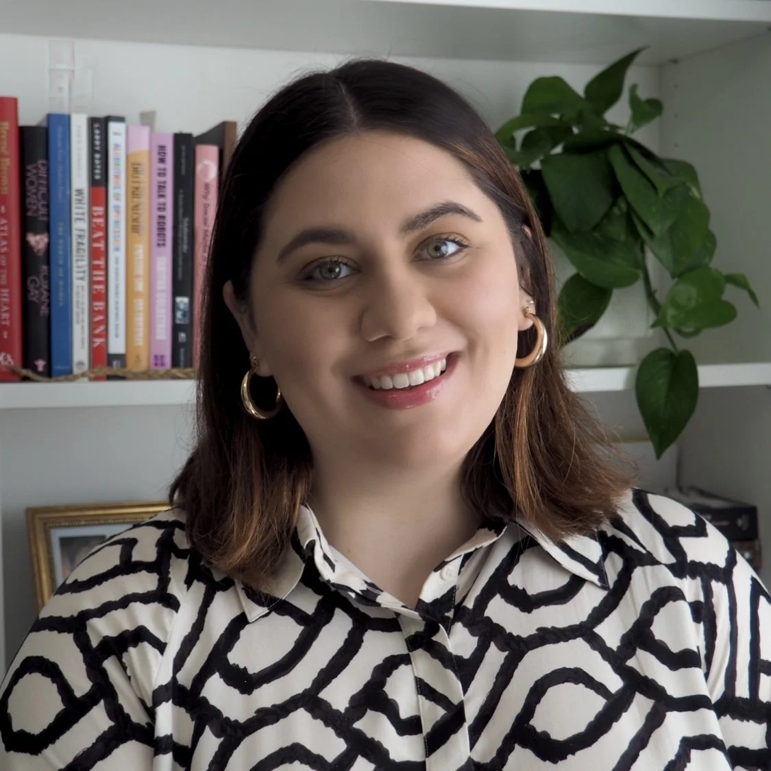 A photo of Alexis-Carlota against a white bookshelf with a plant on the right. She wears a black and white, geometric-pattern shirt with small hoop earrings peeking out from shoulder-length brown hair.