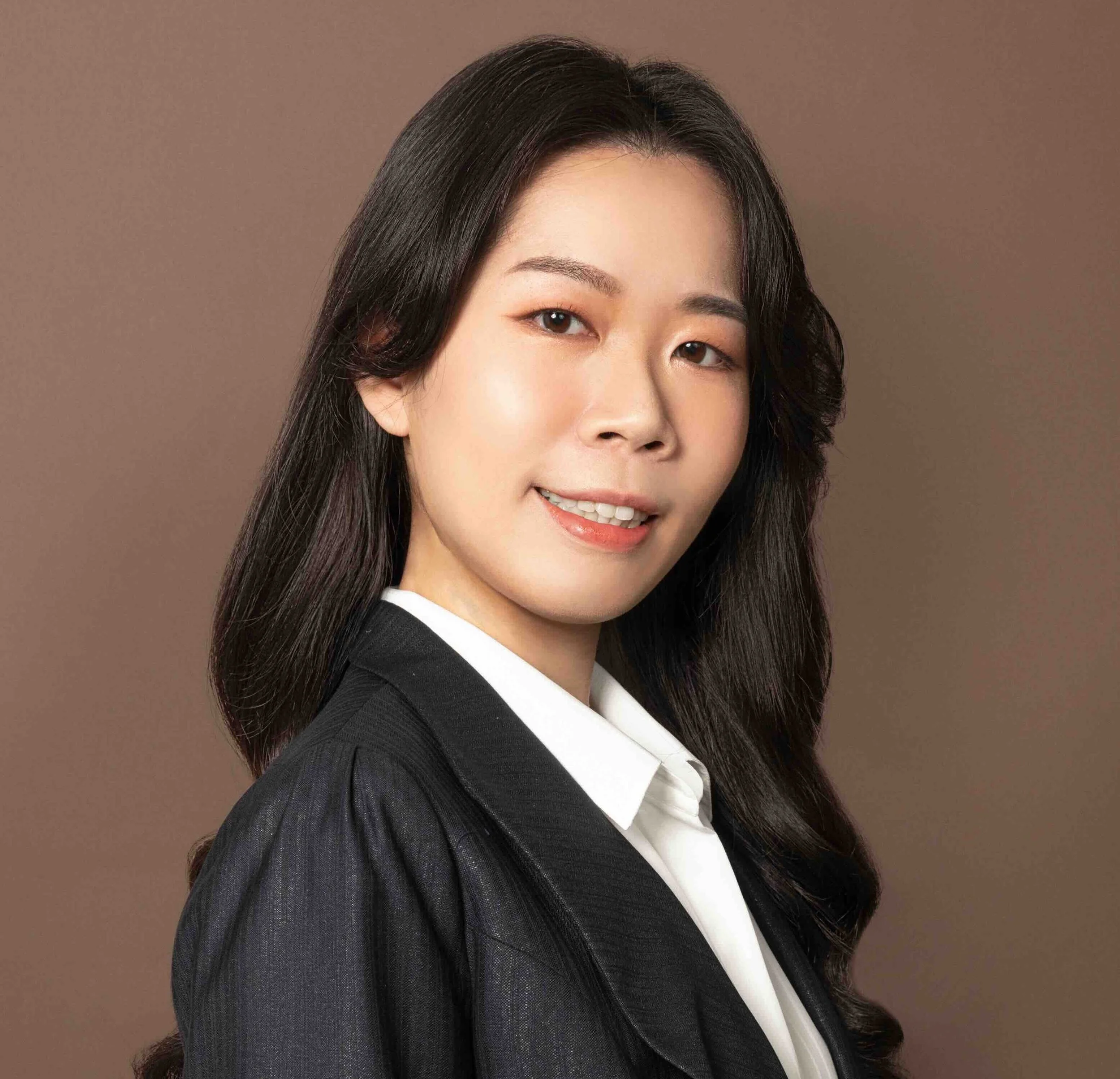 Professional portrait of Cheng-Hsiu, who stands facing the right and looking over her shoulder at the camera. She has long black hair draped over her left shoulder, a kind smile, and stands against a brown background.