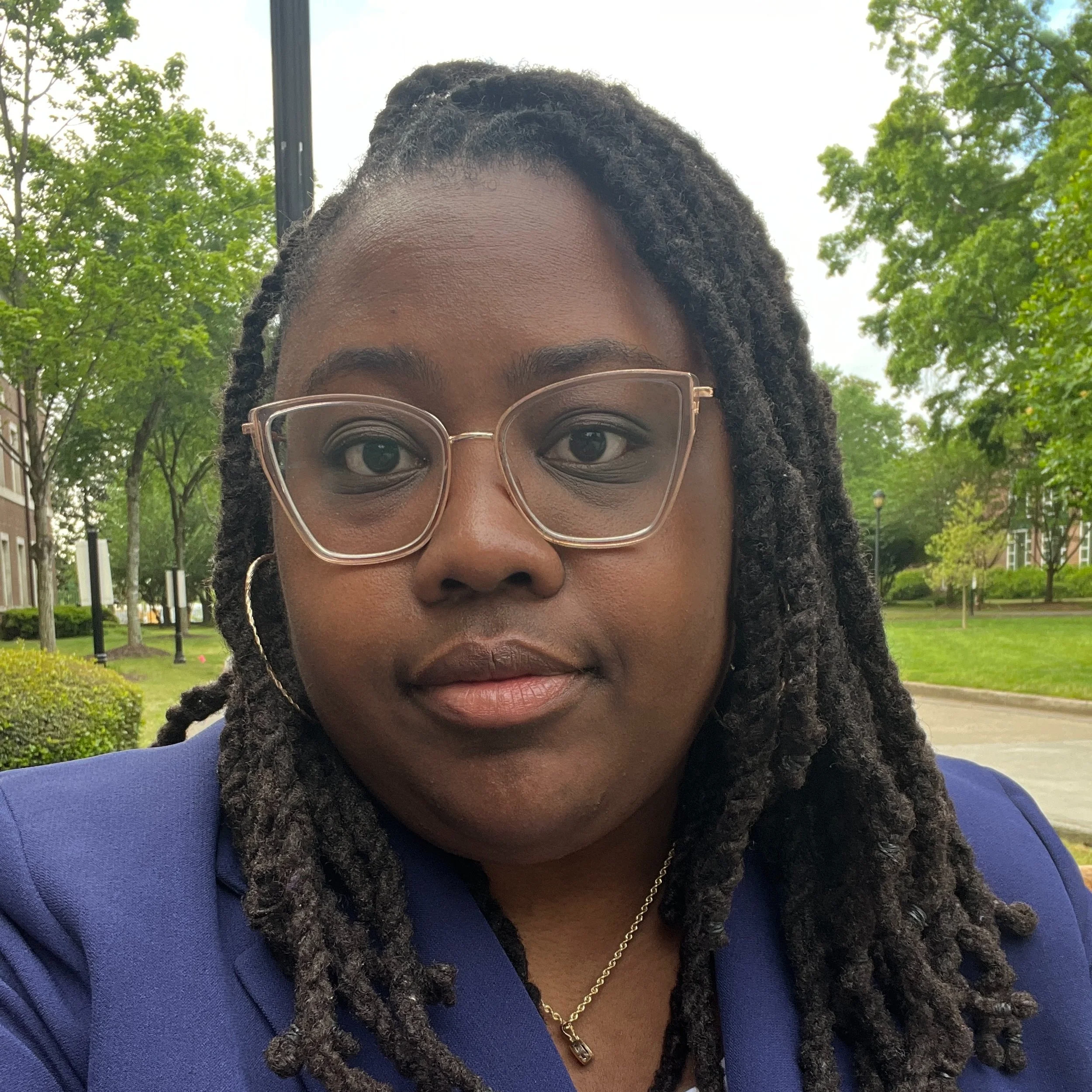 A selfie of Stephanie Jones, a Black woman, outside in a park area. She has thick long braids and wears cat-eye clear plastic glasses, a silver necklace, hoops, and an indigo blazer.