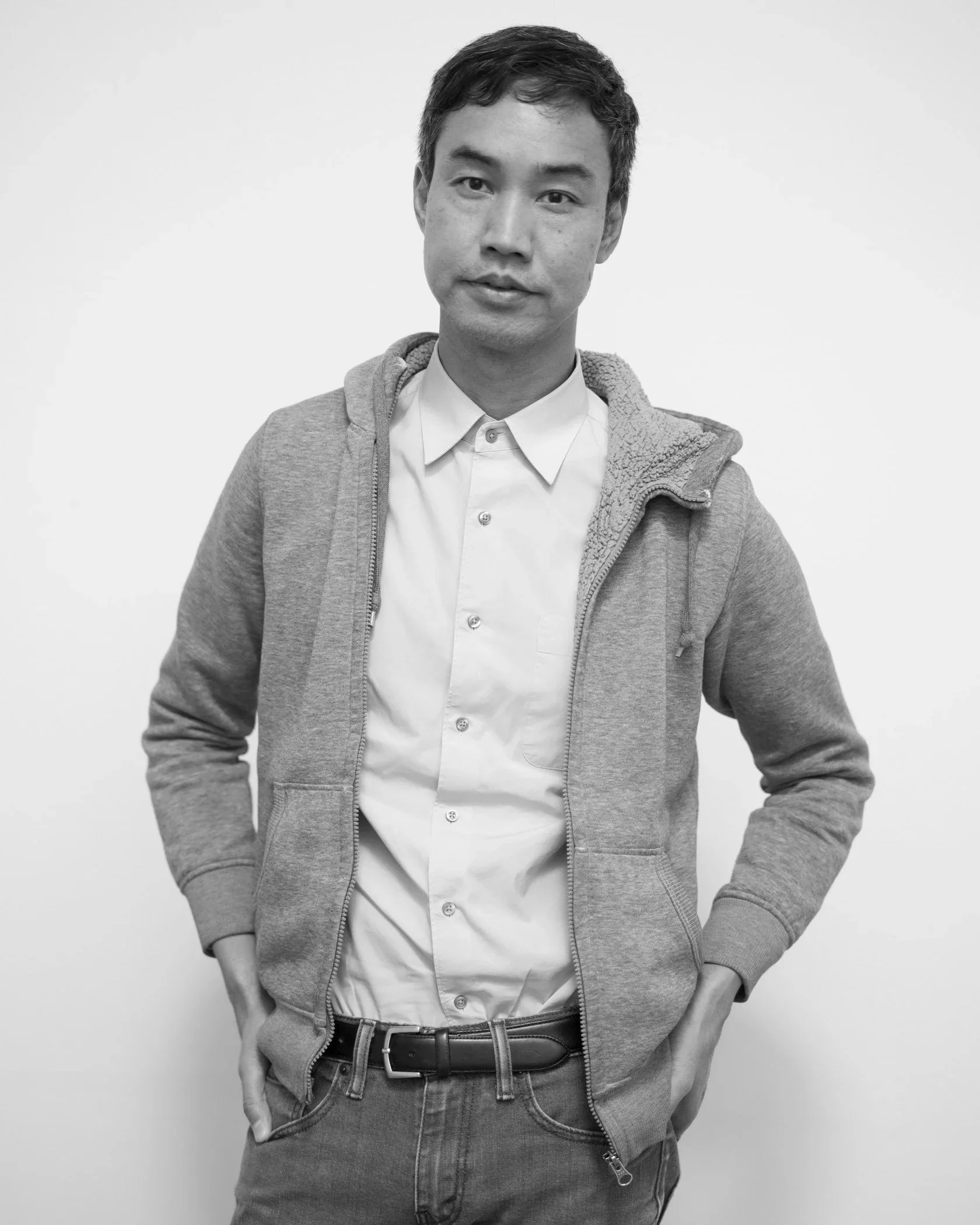 Miljohn is pictured in black and white, with short dark hair, a gray sweater, white button up, and jeans