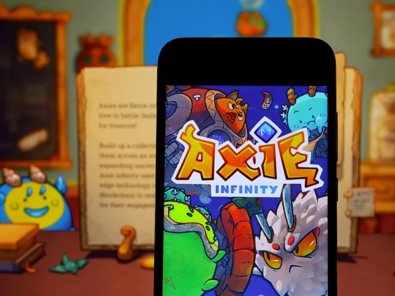 A phone displays a game, Axie Infinity. behind the phone is a blurry cartoon scene