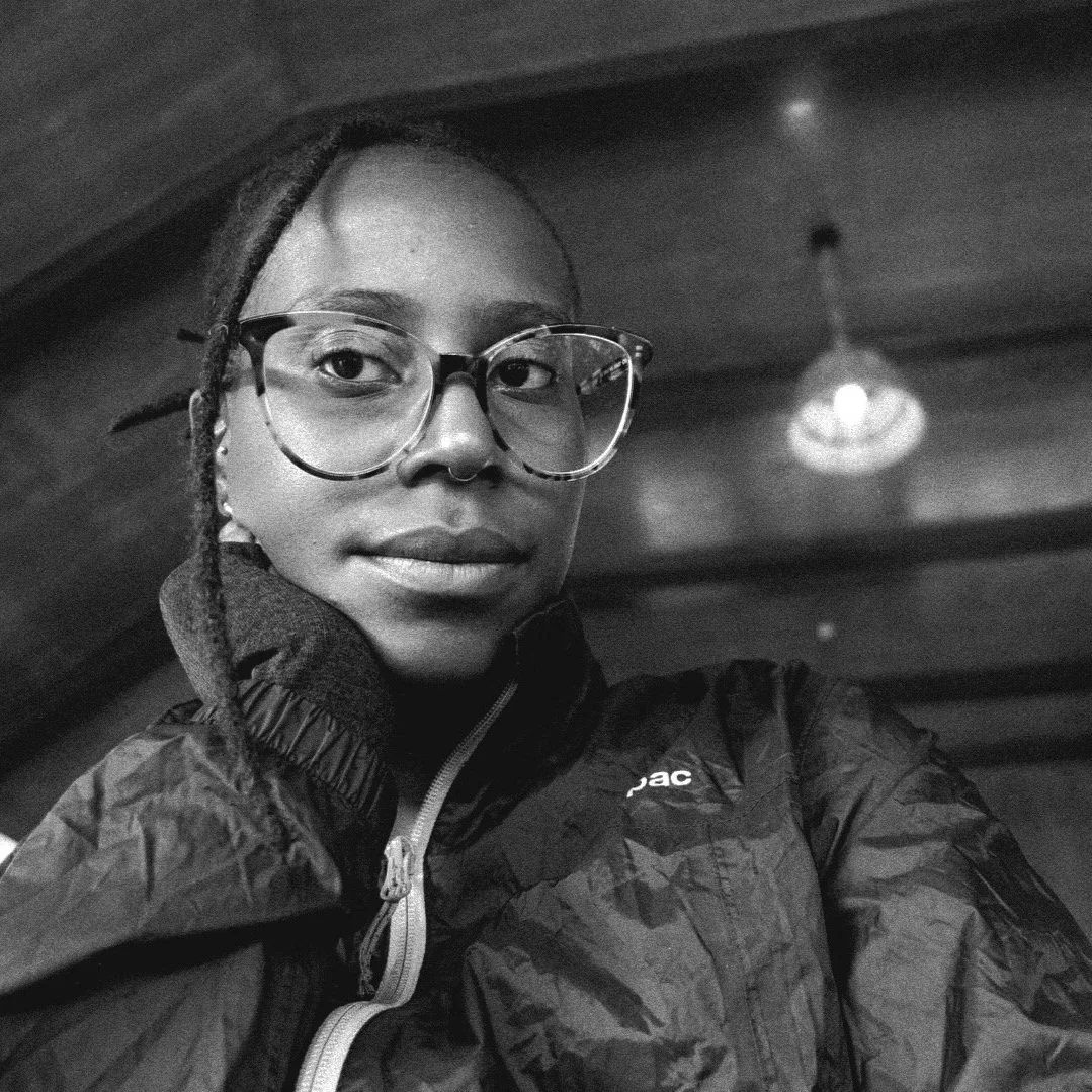 A black and white selfie of tèmí, a Black woman, sitting inside with a ceiling light in the background. She has large tortoise shell glasses, a thin braid framing the left side of her face, and a puffy jacket on.