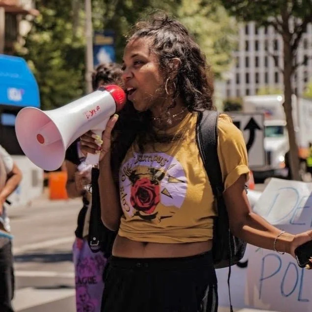 A photo of Vee Copeland, a Brown skinned person with thick, long hair holding a megaphone at a protest. They're mid-sentence, looking to the left, and wear a mustard yellow graphic t-shirt reading "cops off campus."