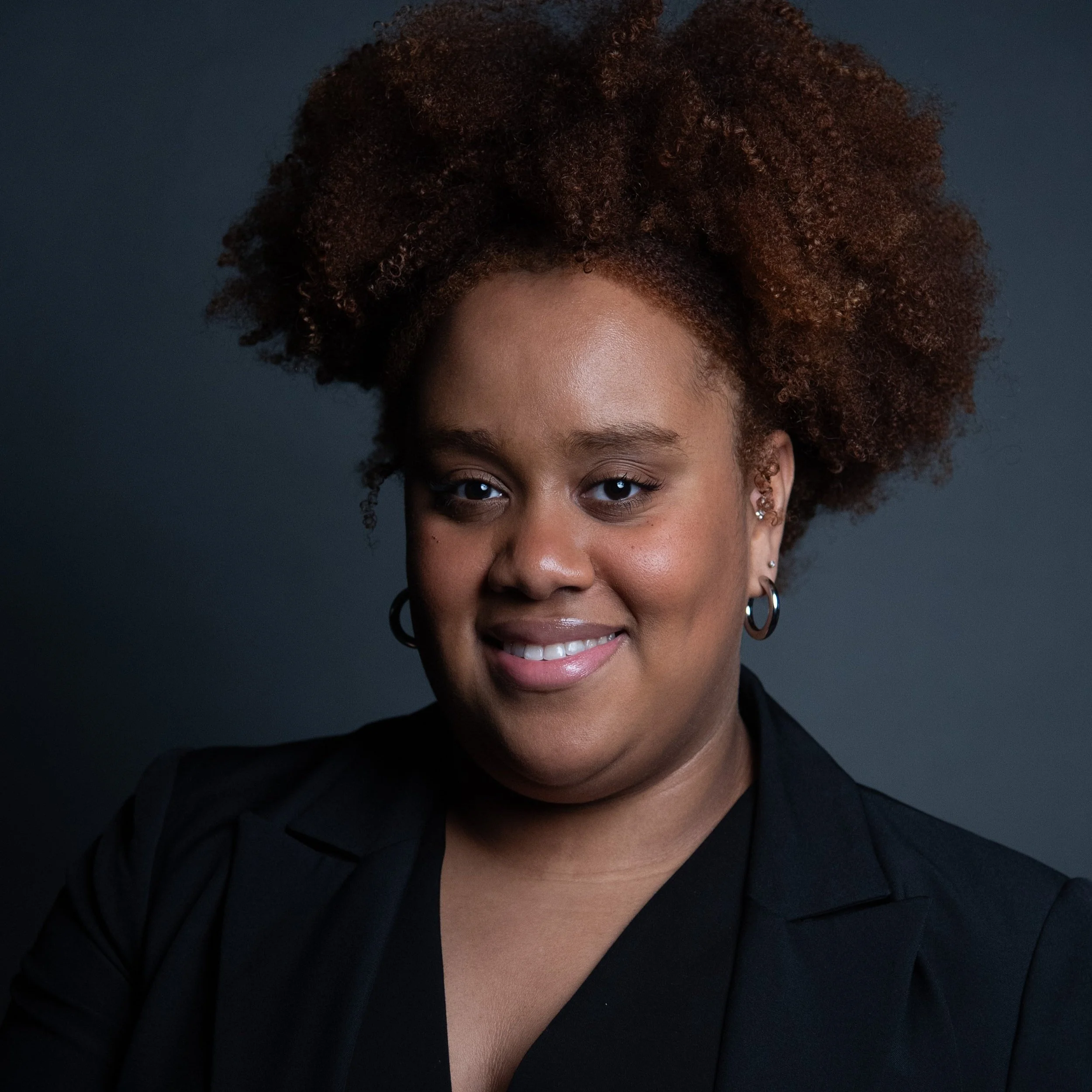 A headshot of Serena Oduro, a Black woman, wearing a black blouse and blazer. She has a sweet smile, wide gauge silver hoop earrings, and a thick, tall afro hairstyle.