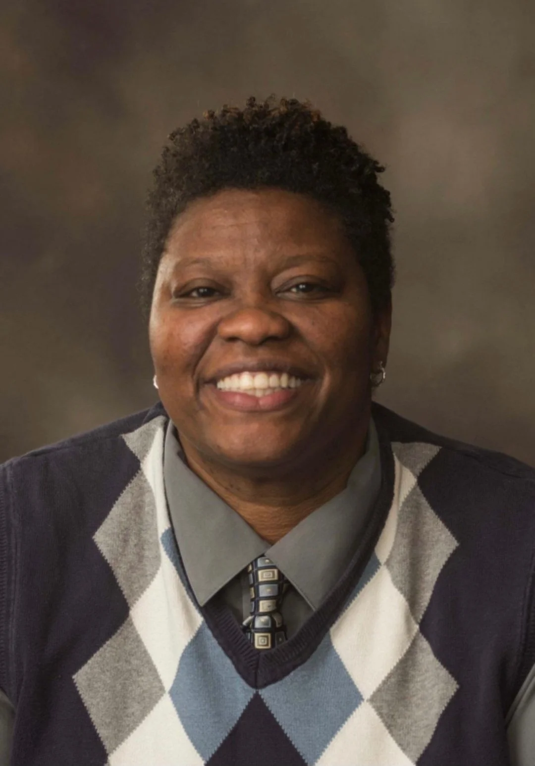 A headshot of Chris Wiley, a Black man, sitting against a gray backdrop. He has shorter hair, deep brown eyes, a diamond pattern sweater, small silver hoop earrings, and a wide smile.