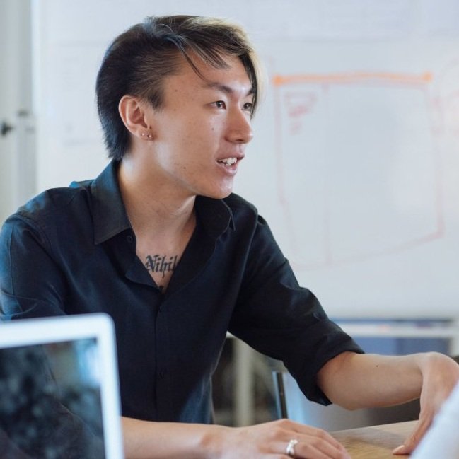 A photo of Wells Santo, an East Asian nonbinary person, sitting in an office space talking to someone. She wears a black button down, a silver ring, two small stud earrings, and has a partly visible chest tattoo with the letters "Nihil."