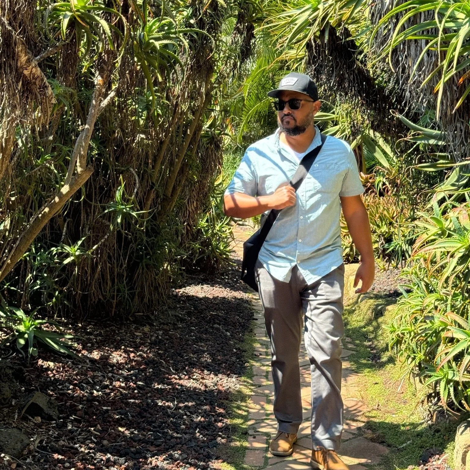 A photo of Beza Merid, a Black man, walking outside on a brick path between tall tropical plants. He wears a baseball hat, sunglasses, a pale blue button down, and grips his messenger bag.