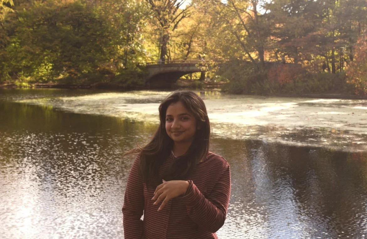 Photo of Xeneb Shah, a South Asian woman wearing a striped shirt posing outdoors at a park with autumn leaves.