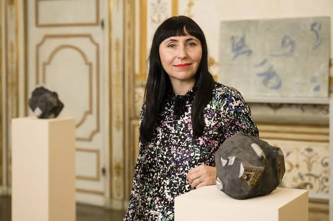 Agnieszka Kurant has dark straight hair, a colorful blouse, and poses beside a sculpture