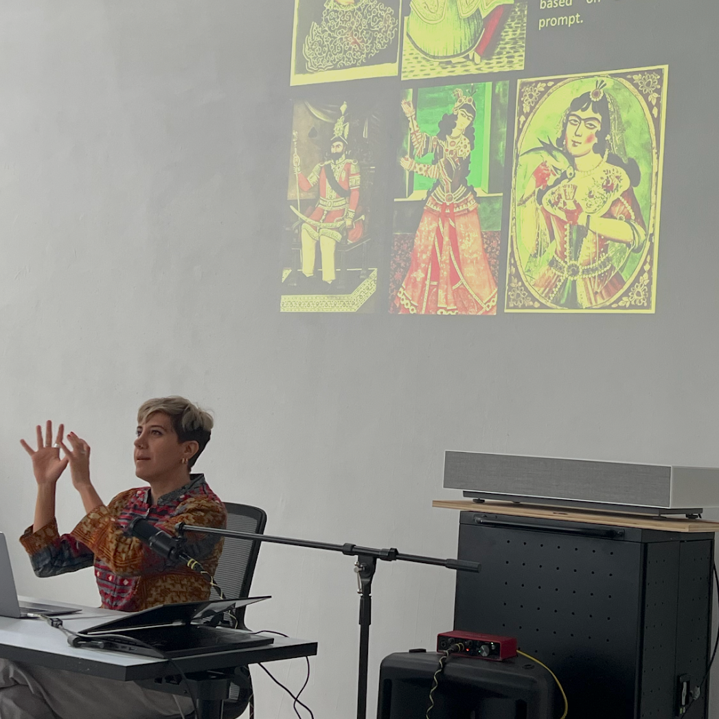 A photo of Morehshin Allahyari giving a talk, sat at a desk with a microphone and a laptop, a yellow projection of saint images on the wall behind