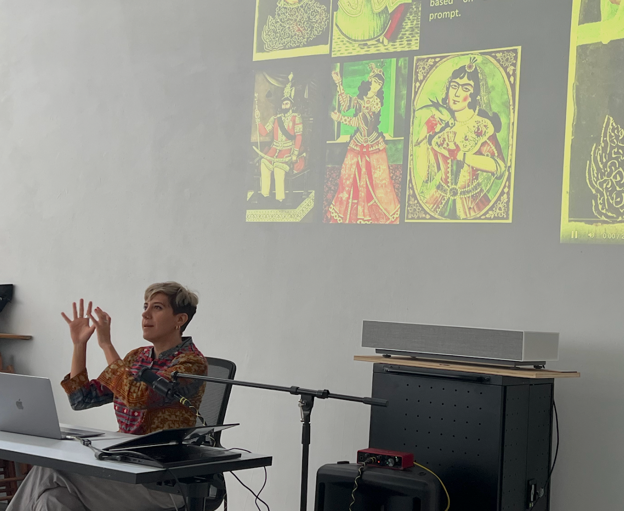 A photo of Morehshin Allahyari giving a talk, sat at a desk with a microphone and a laptop, a yellow projection of saint images on the wall behind