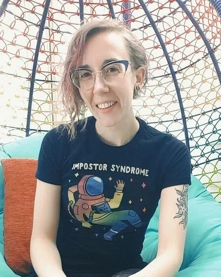 A photo of Remi Yergeau, a white person with a graphic t shirt, glasses, and hair hanging beside their face
