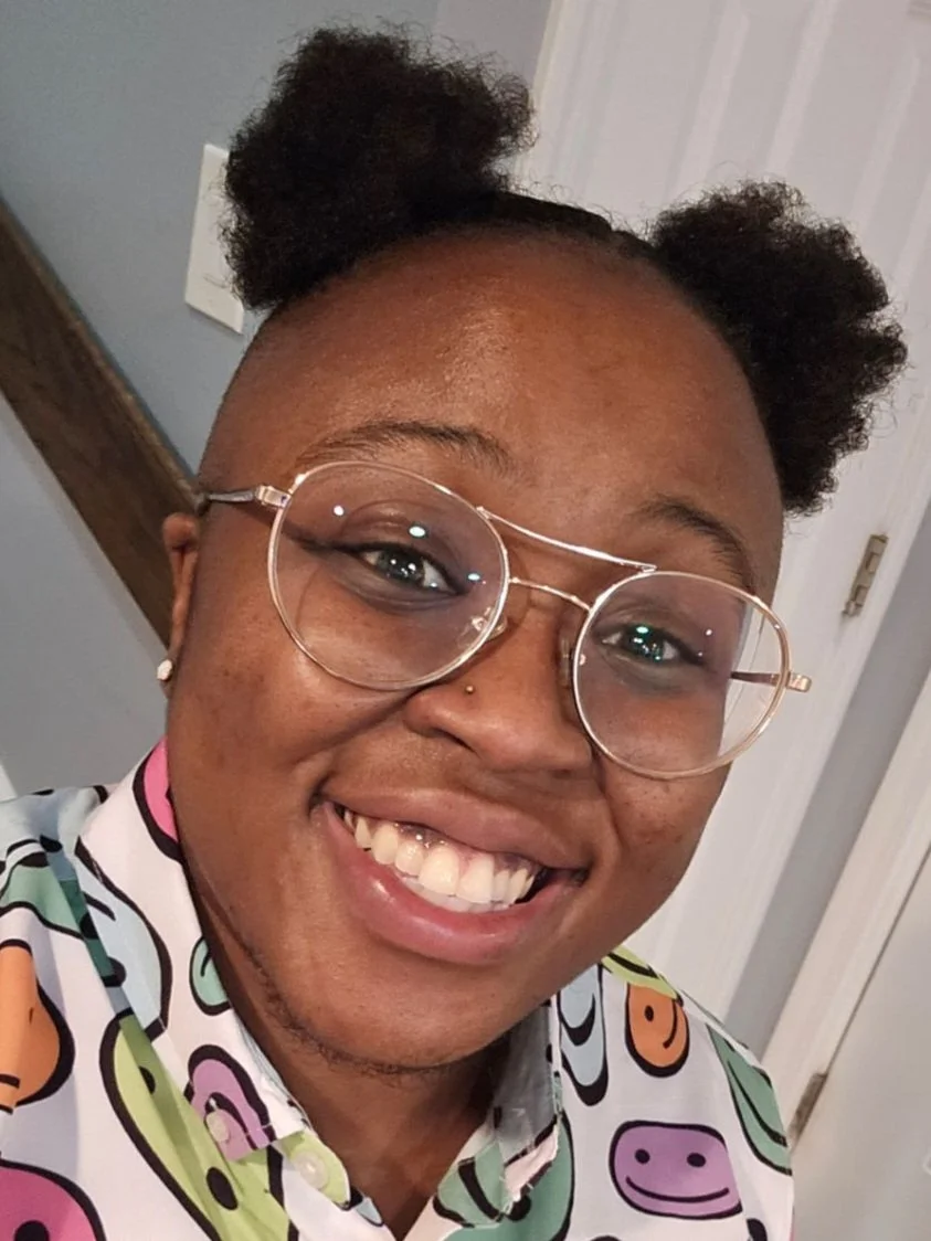 Headshot of Dez Brown, a Black nonbinary person with wire-frame glasses and a wide smile.