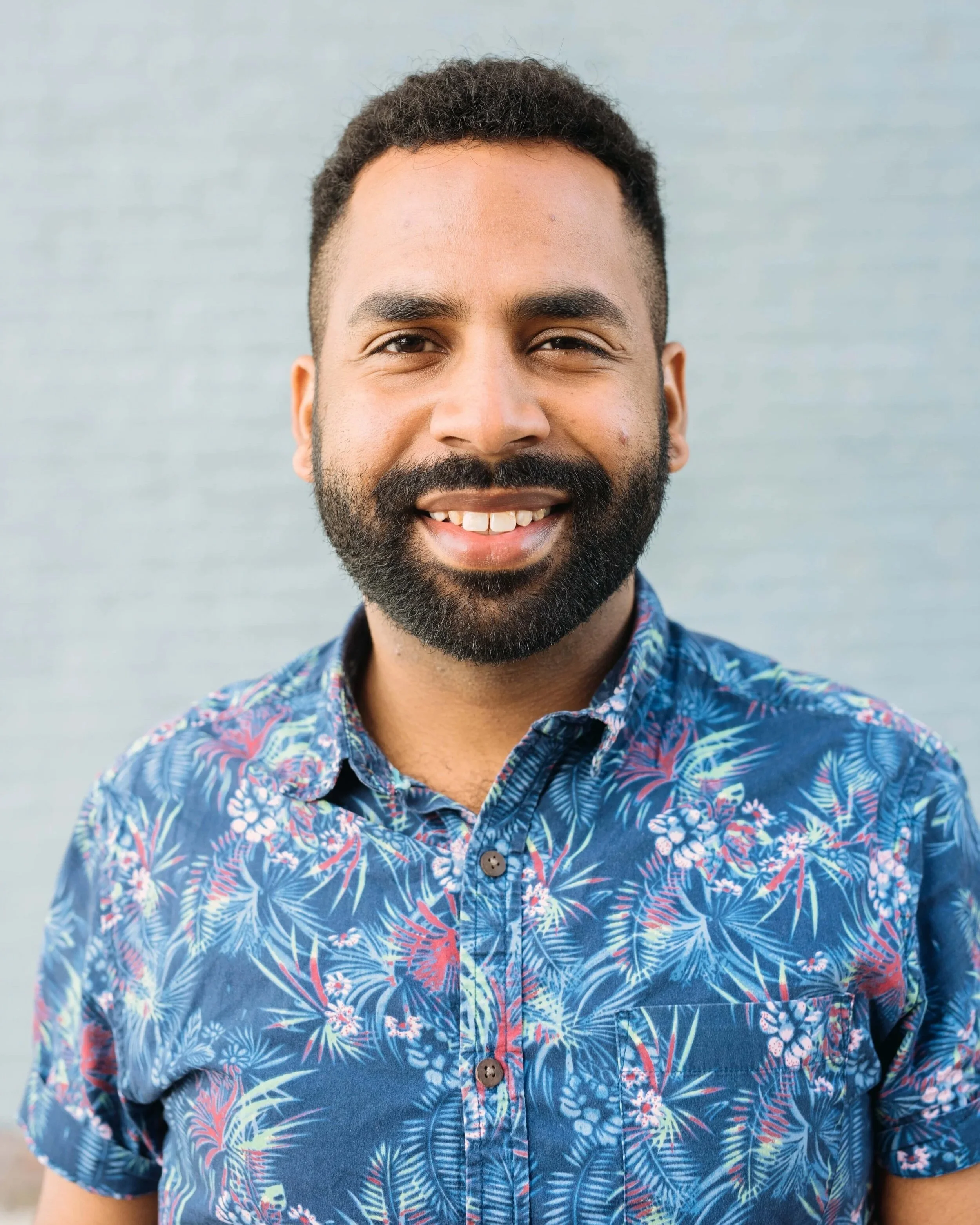 Johann has brown skin, a dark beard and hair, a smile, a blue aloha pattern shirt