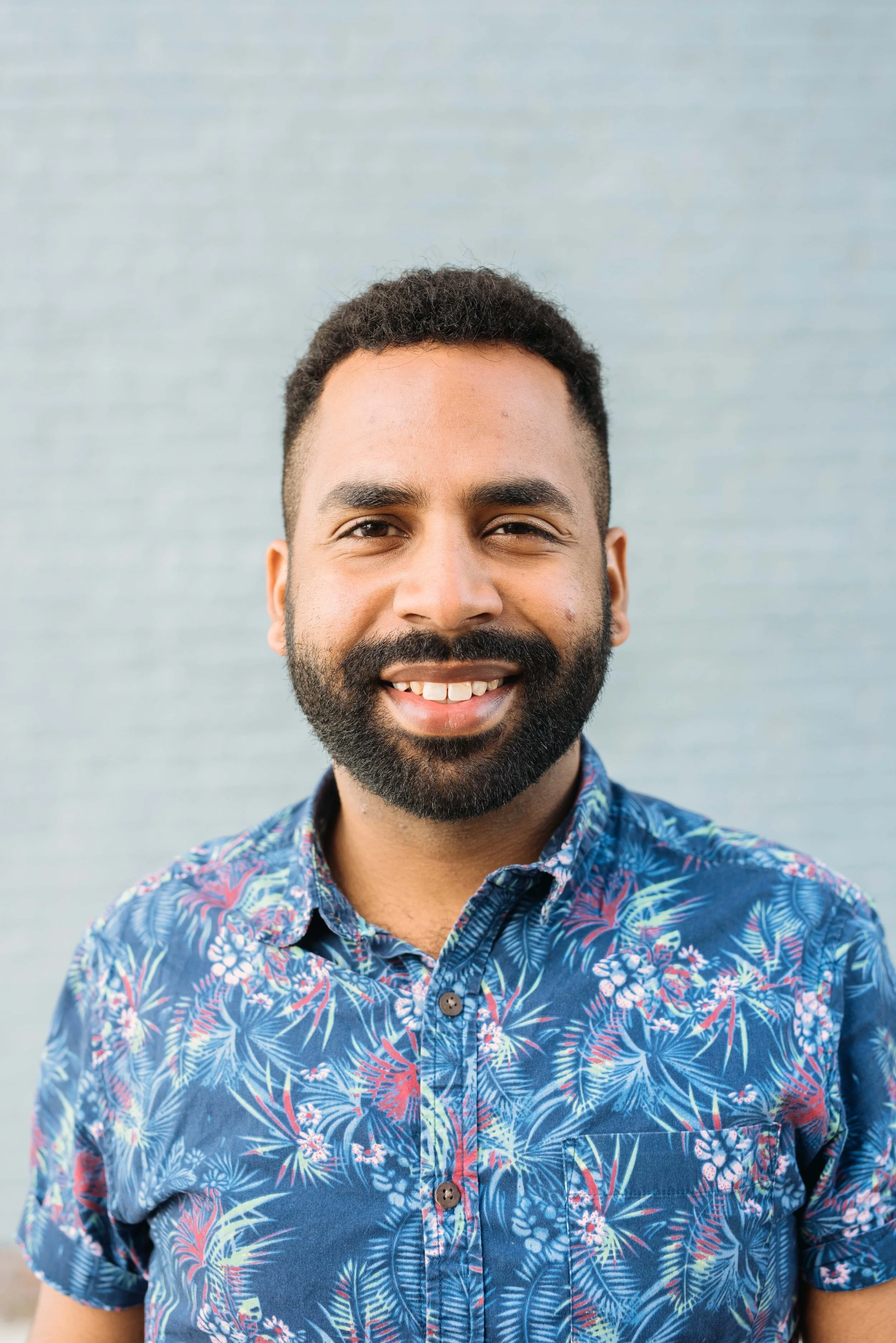 Johann has brown skin, a dark beard and hair, a smile, a blue aloha pattern shirt