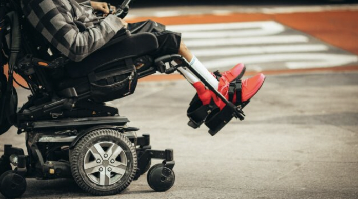 the bottom half of a power wheelchair, with someone's legs and feet strapped in. The person is wearing bright red shoes and white socks.