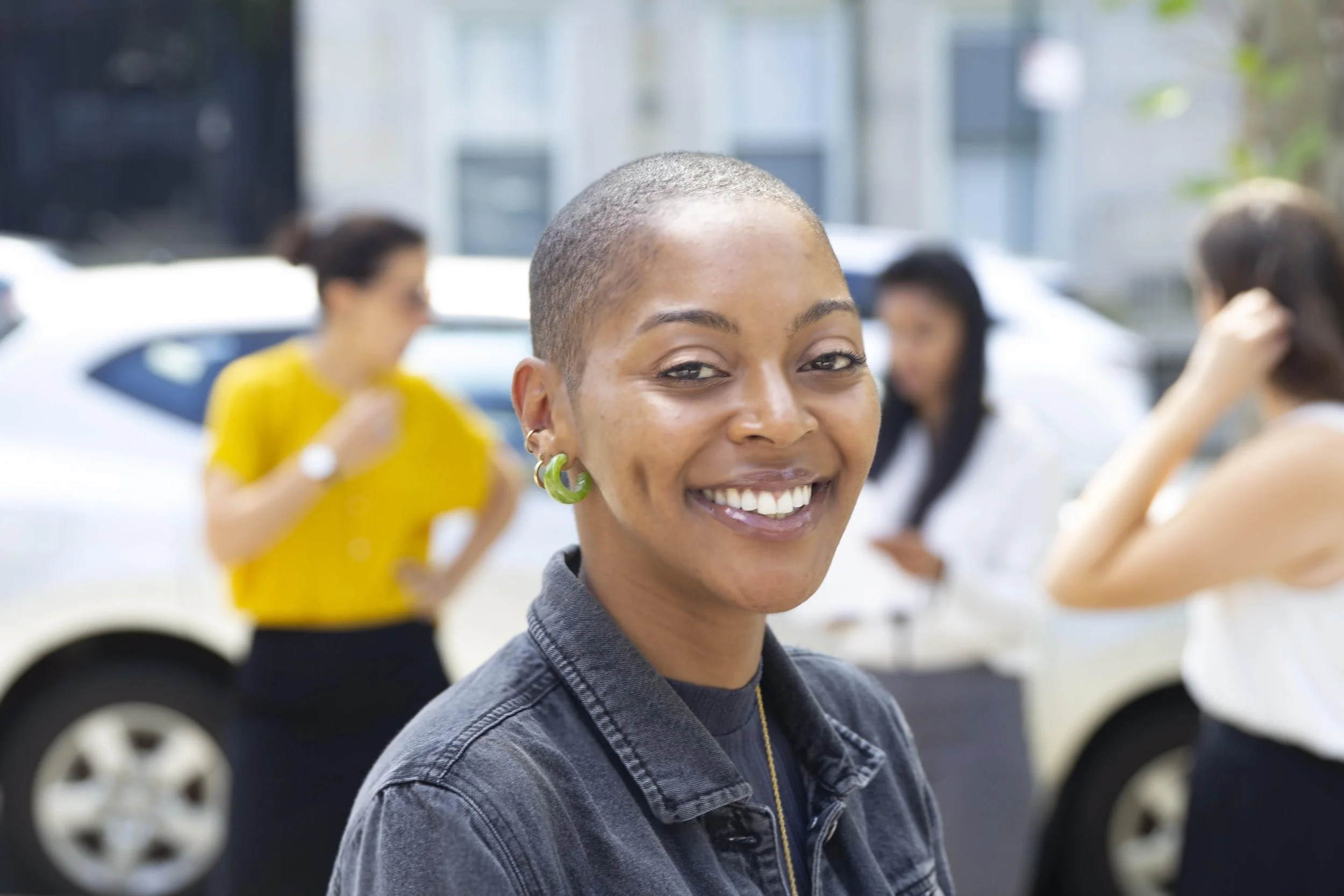 A headshot of Chelsea Allen, a Black woman with a shaved hairstyle wearing a denim jacket and large green earrings.